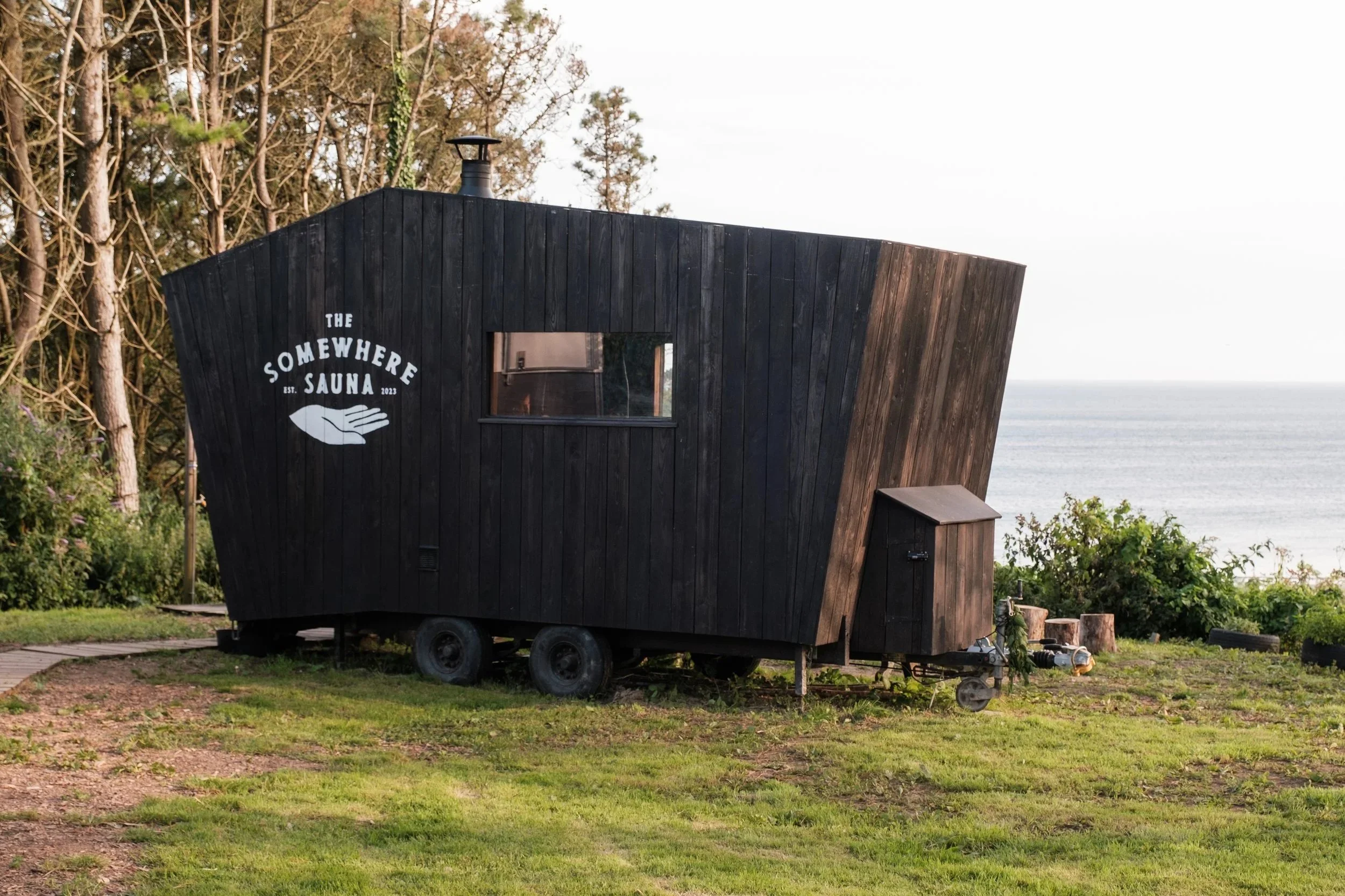 A black wooden tiny house on wheels with a sign that reads 'The Somewhere Sauna,' located outdoors near the water, with trees and grass surrounding it.