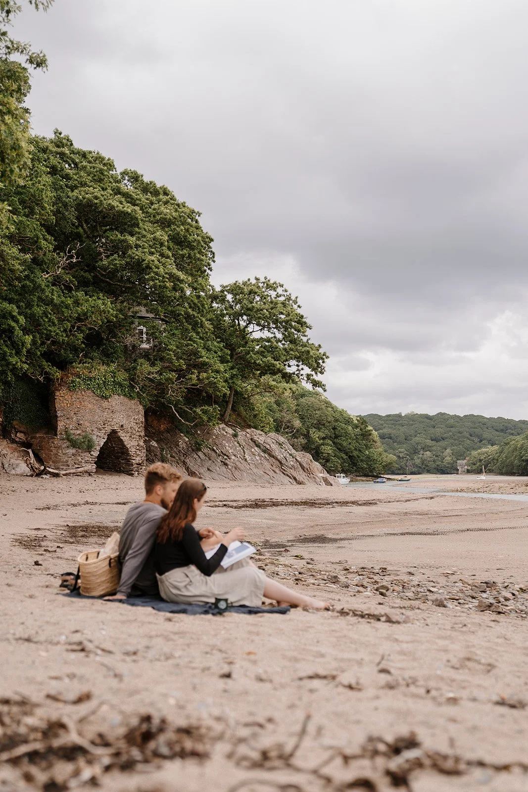Couple sitting on a sandy beach, reading a book under a cloudy sky. Trees and rocky cliffs in the background.