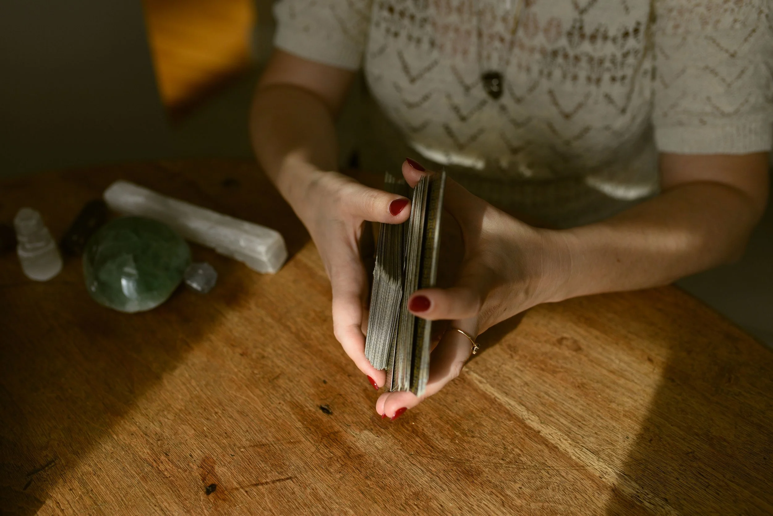 A person with red nail polish and a ring on finger holding a deck of tarot cards over a wooden table with various crystals and gemstones.