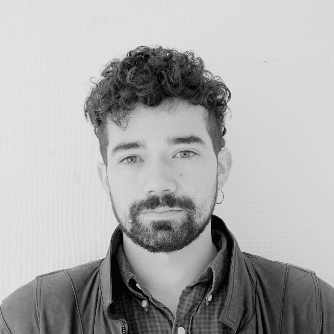 A black-and-white portrait of a young man with curly hair, a beard, and light-colored eyes, wearing a leather jacket over a button-up shirt, standing against a plain background.