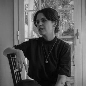 Black-and-white photo of a woman sitting at a table near a window, wearing a dark top and a necklace.