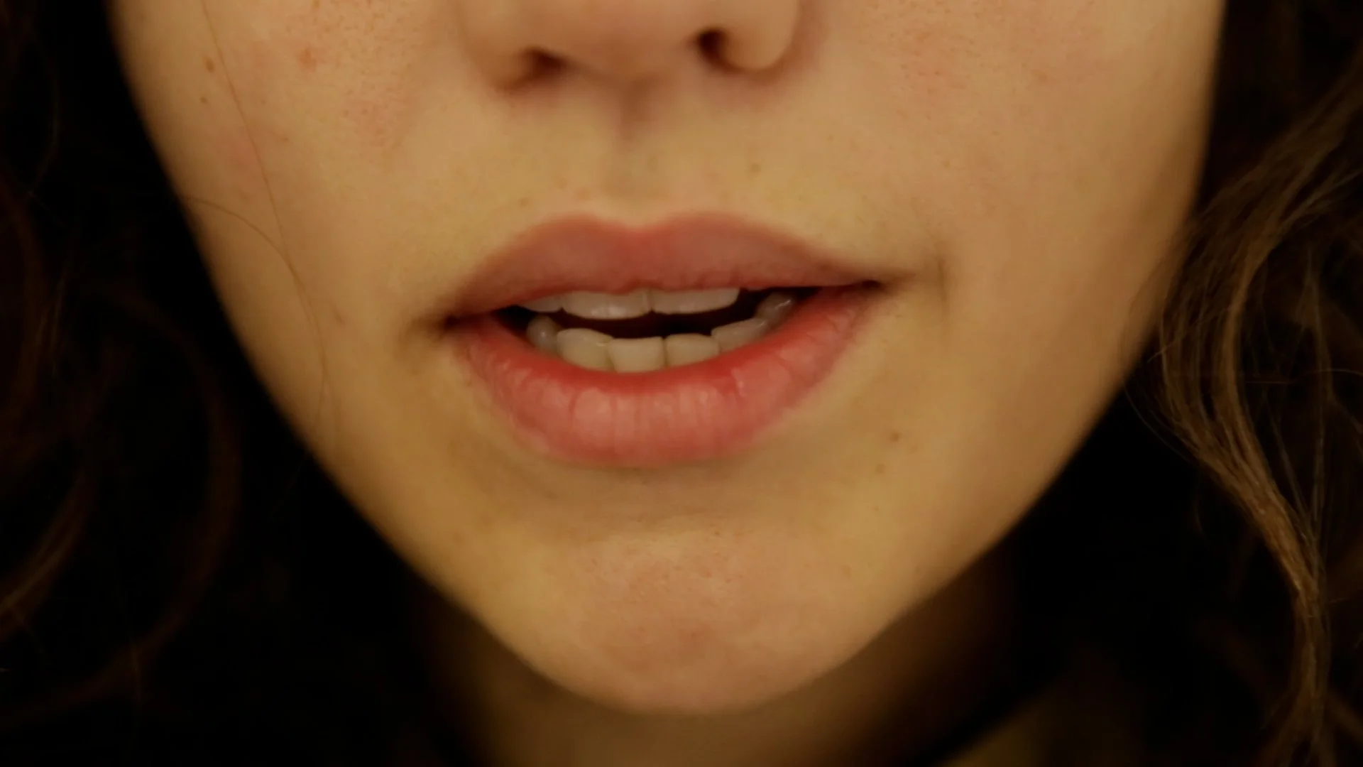 Close-up of a person's face with lips slightly parted, showing teeth and tongue, with visible freckles and brown hair.