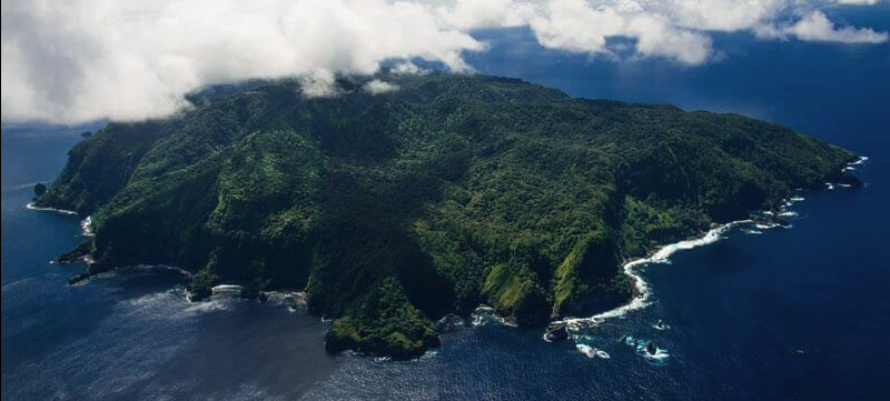 An aerial view of a lush green island surrounded by ocean with waves crashing against the shore, partly covered by clouds.