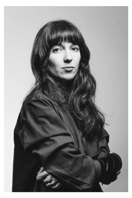 Black and white portrait of a woman with long wavy hair and straight bangs, wearing a dark jacket and gloves, with arms crossed, looking at the camera.