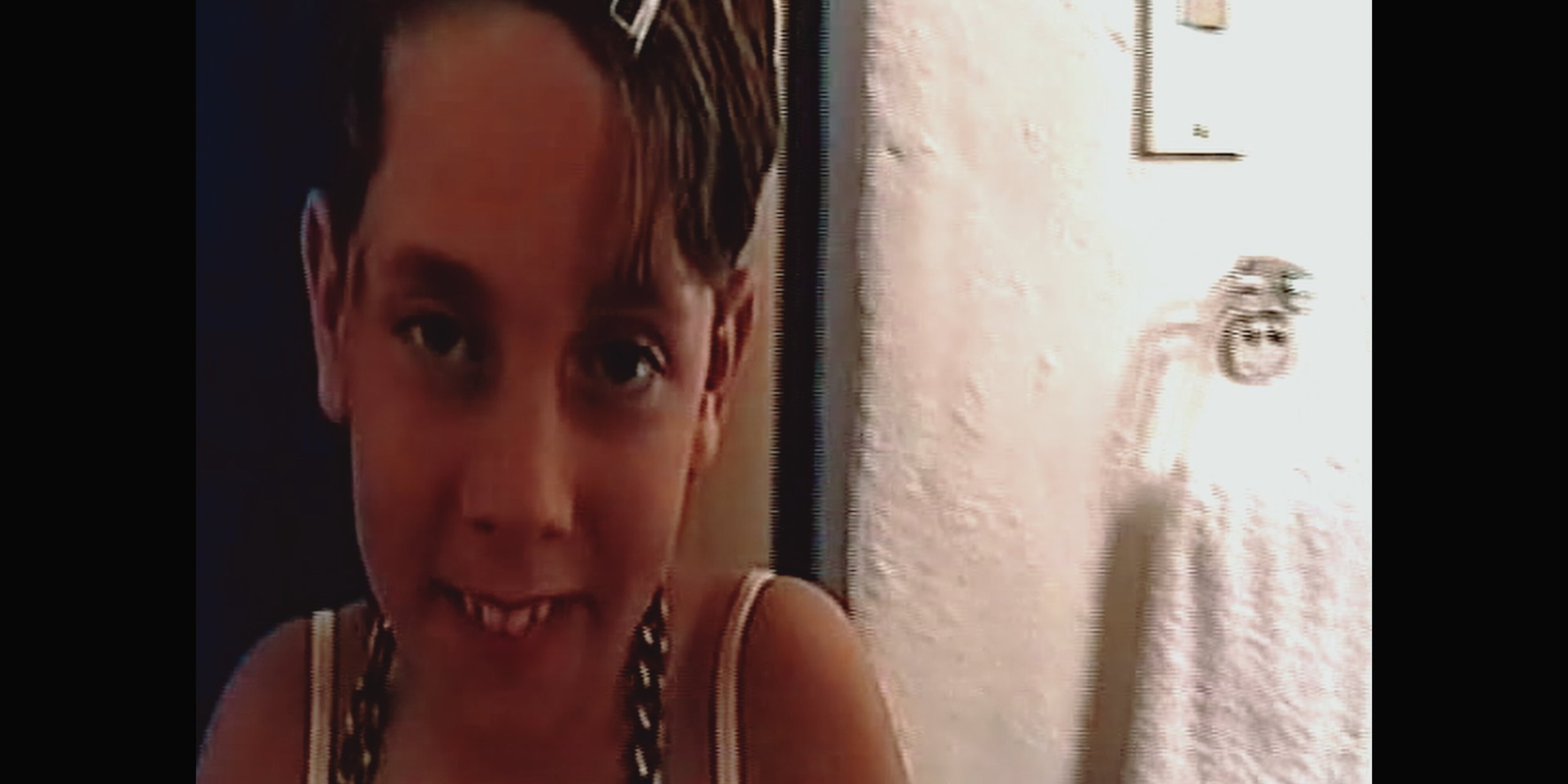 A young girl with brown hair, smiling with teeth visible, wearing a striped tank top, inside a room with a white wall and a framed picture.