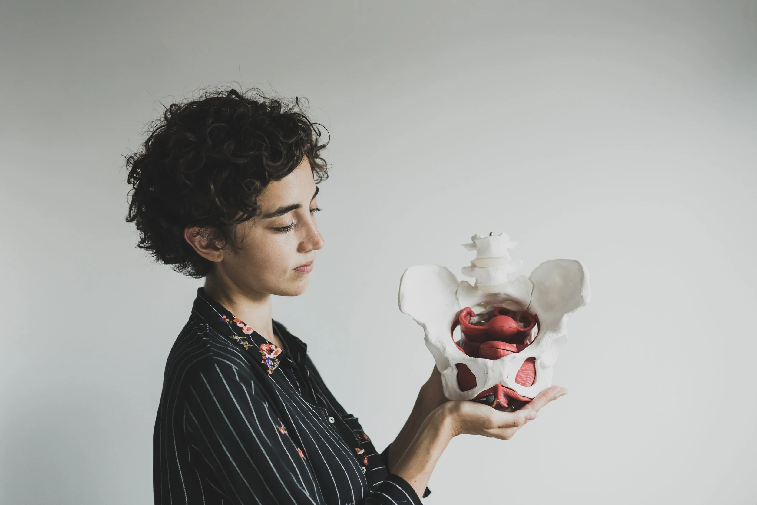 A woman with short curly dark hair looking at a human pelvic bone model she is holding.