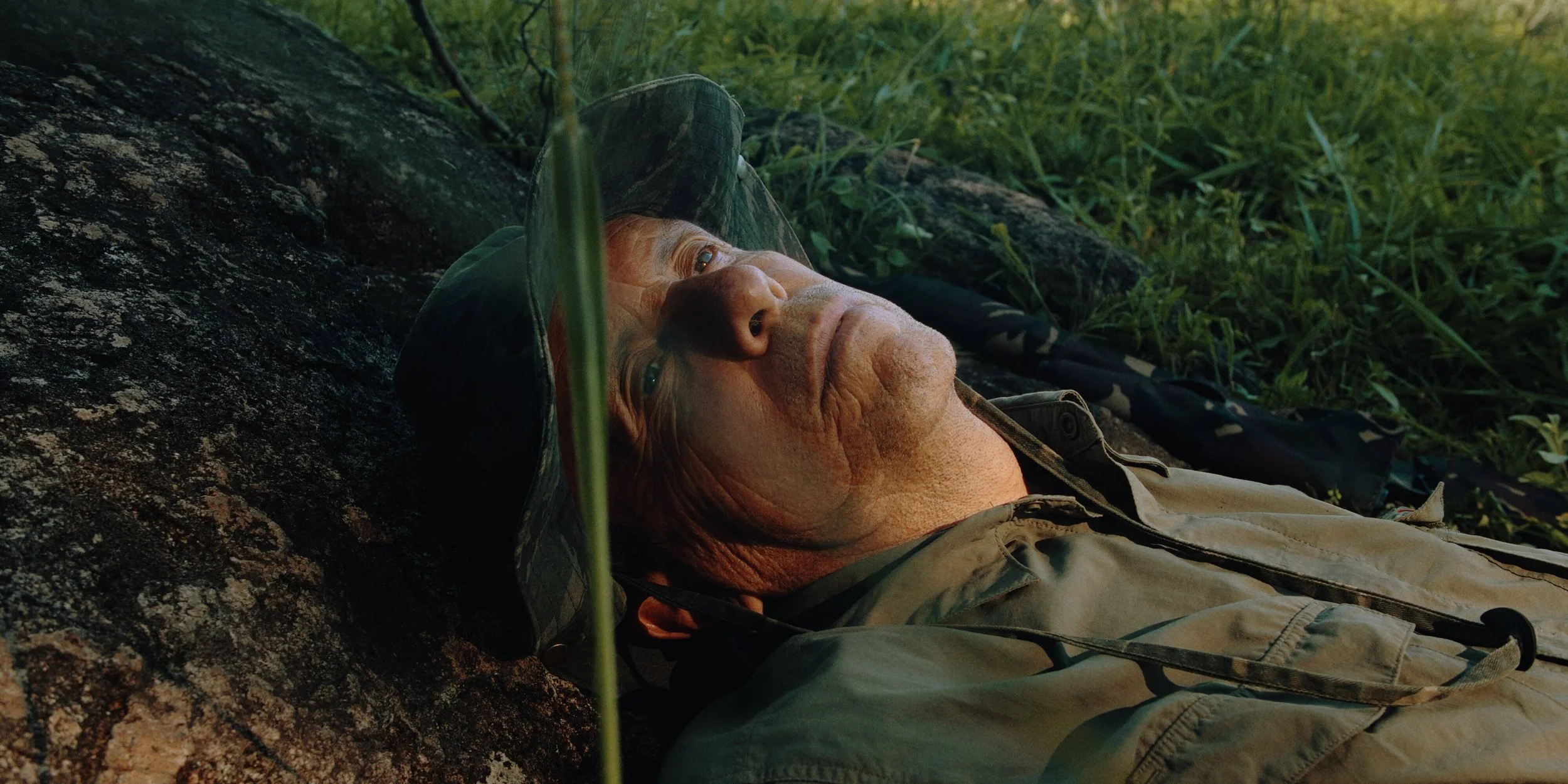 A person lying on the ground in a natural outdoor setting, wearing a wide-brimmed hat and camouflage clothing, looking up with a calm expression.