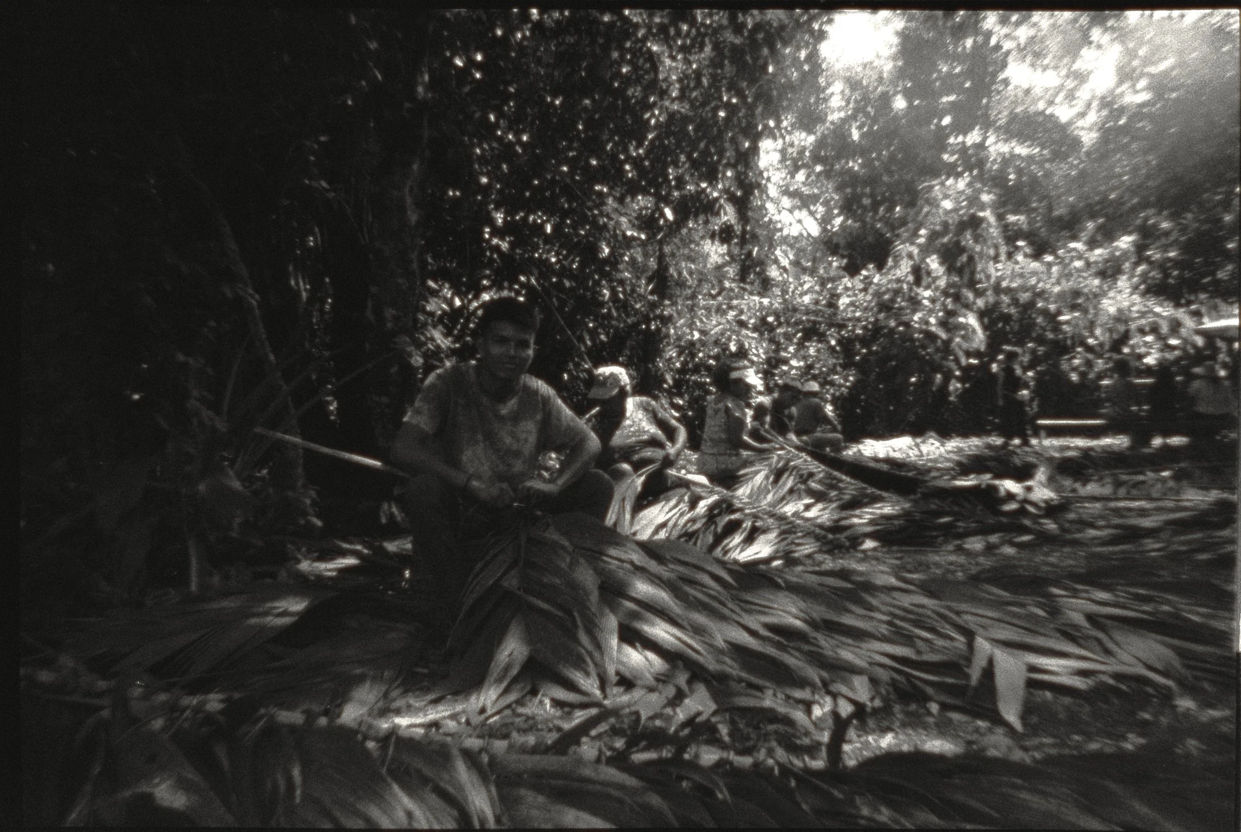 A black and white photo of several children sitting on the ground surrounded by dense foliage.