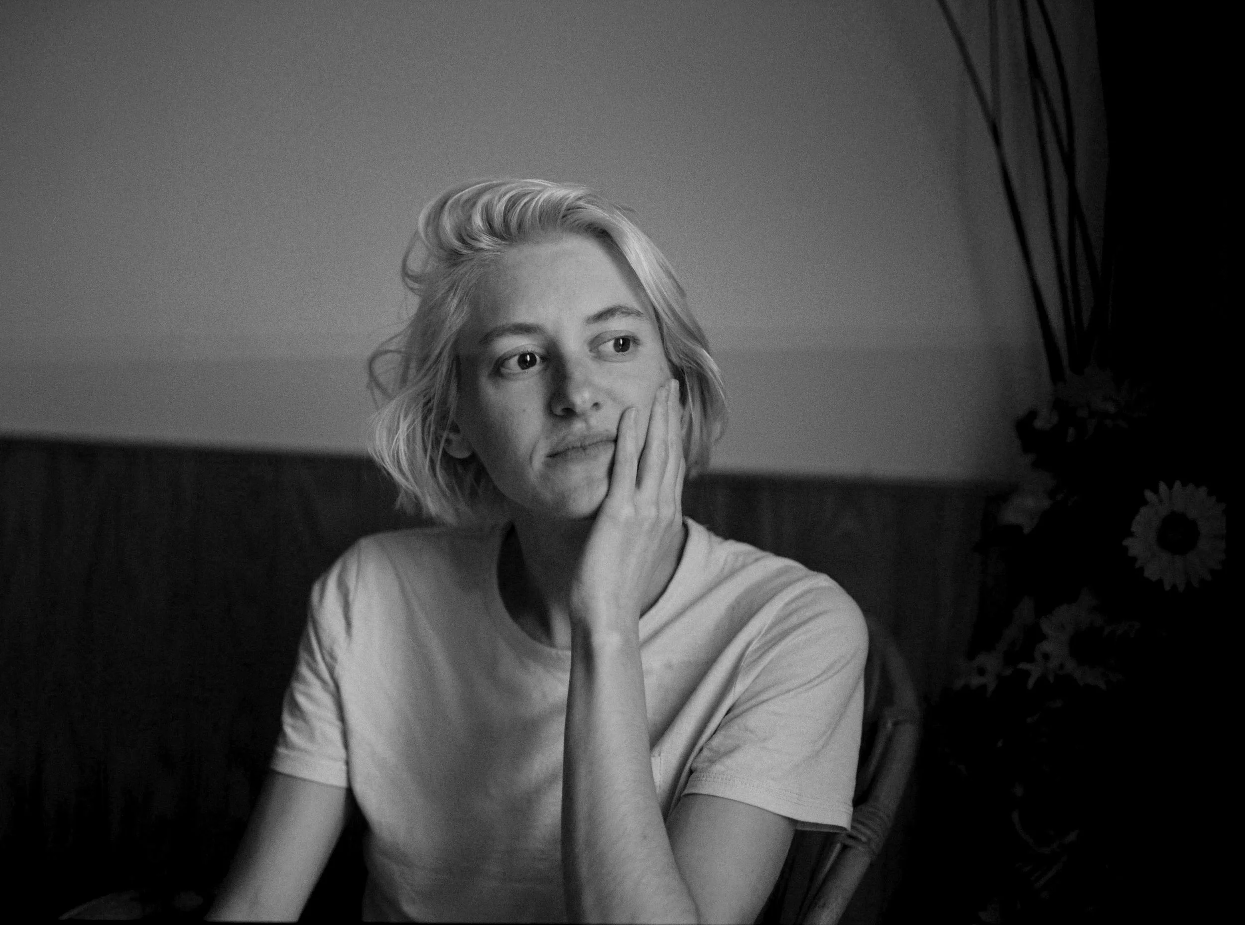 A black and white photo of a young woman with short, wavy hair, wearing a light-colored t-shirt, resting her chin on her hand and looking to the side with a thoughtful expression.