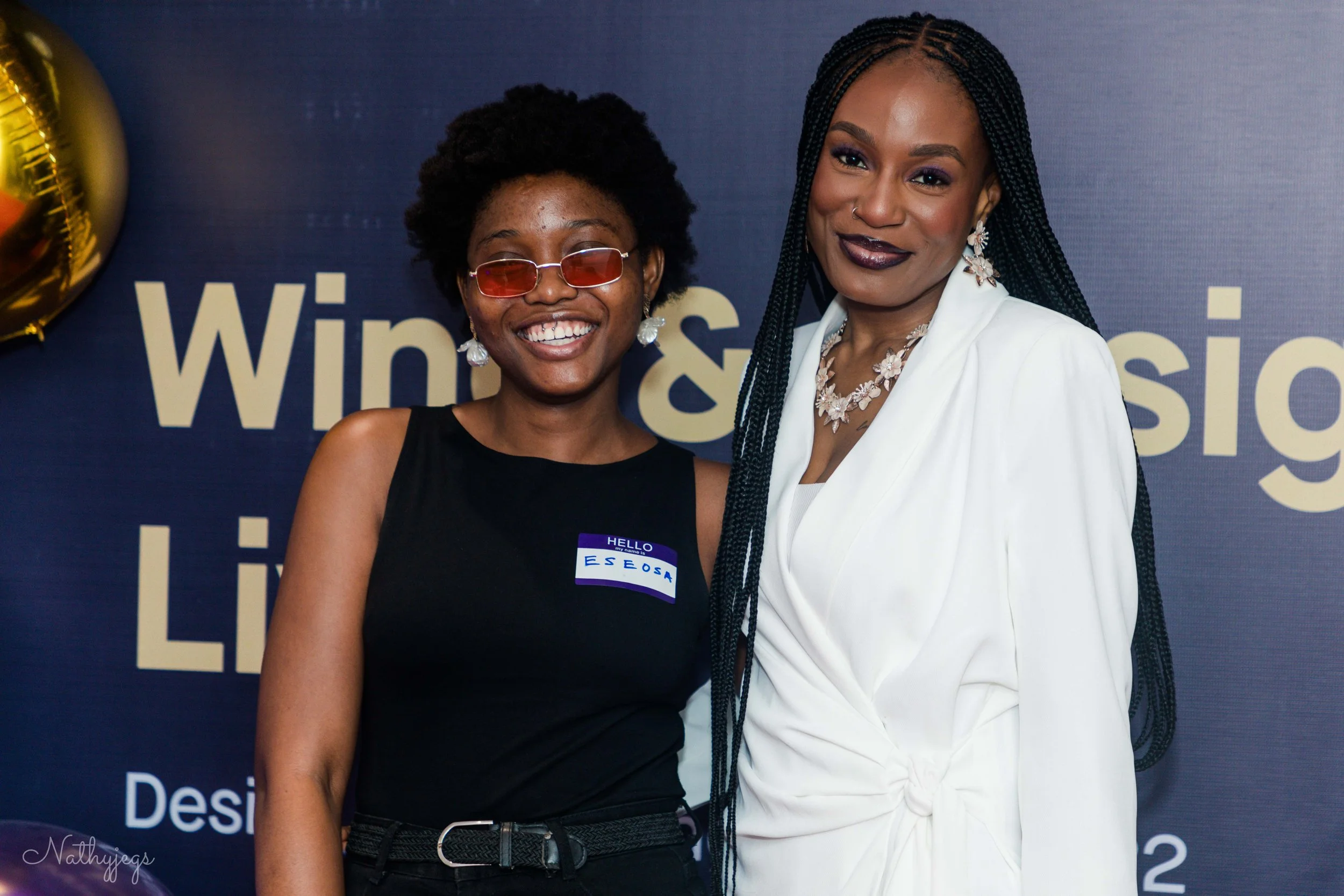 Two women standing together at an event, smiling. One woman wears sunglasses and a black sleeveless top with a name tag. The other woman wears a white blazer and jewelry. A dark background with text and a balloon is partially visible.