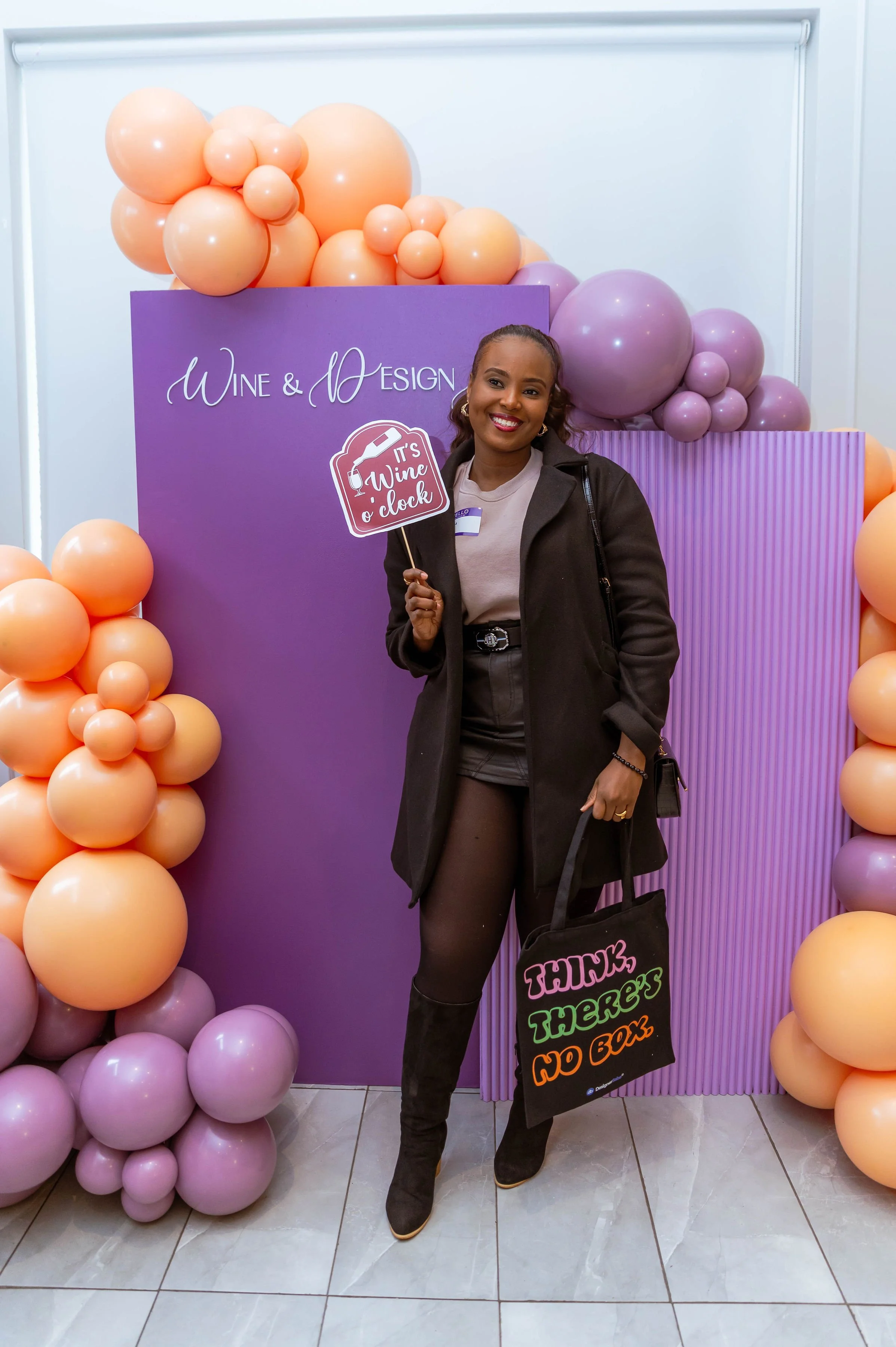 A woman smiling at a wine and design event, holding a sign that reads 'It's Wine o' clock'. She is standing in front of a purple decorative backdrop surrounded by pink and peach-colored balloons, wearing black boots, black shorts, a beige top, and a 