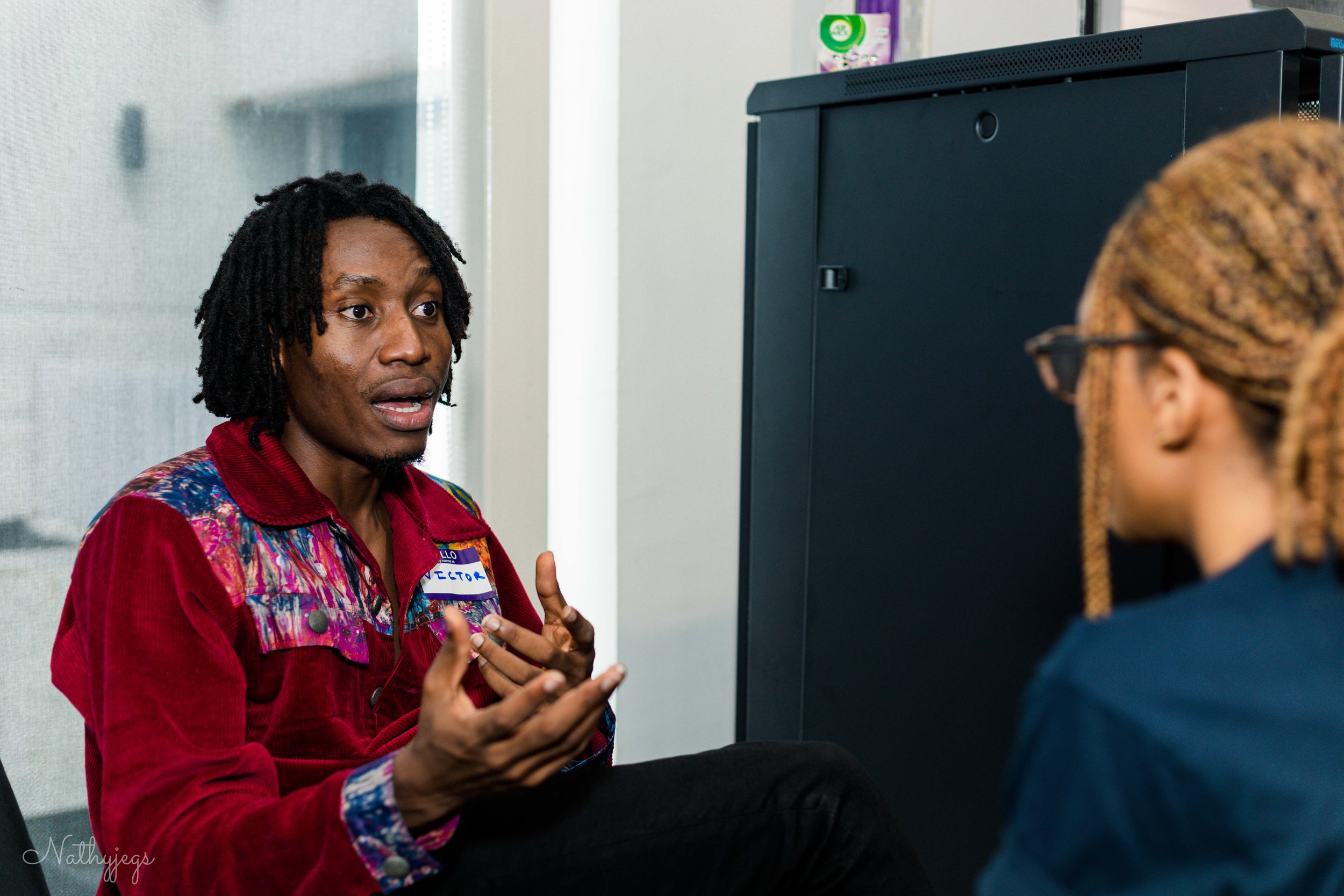 Two people engaged in a serious conversation, one wearing a red jacket with a name tag, and the other with glasses and braided hair, indoors near a black cabinet.