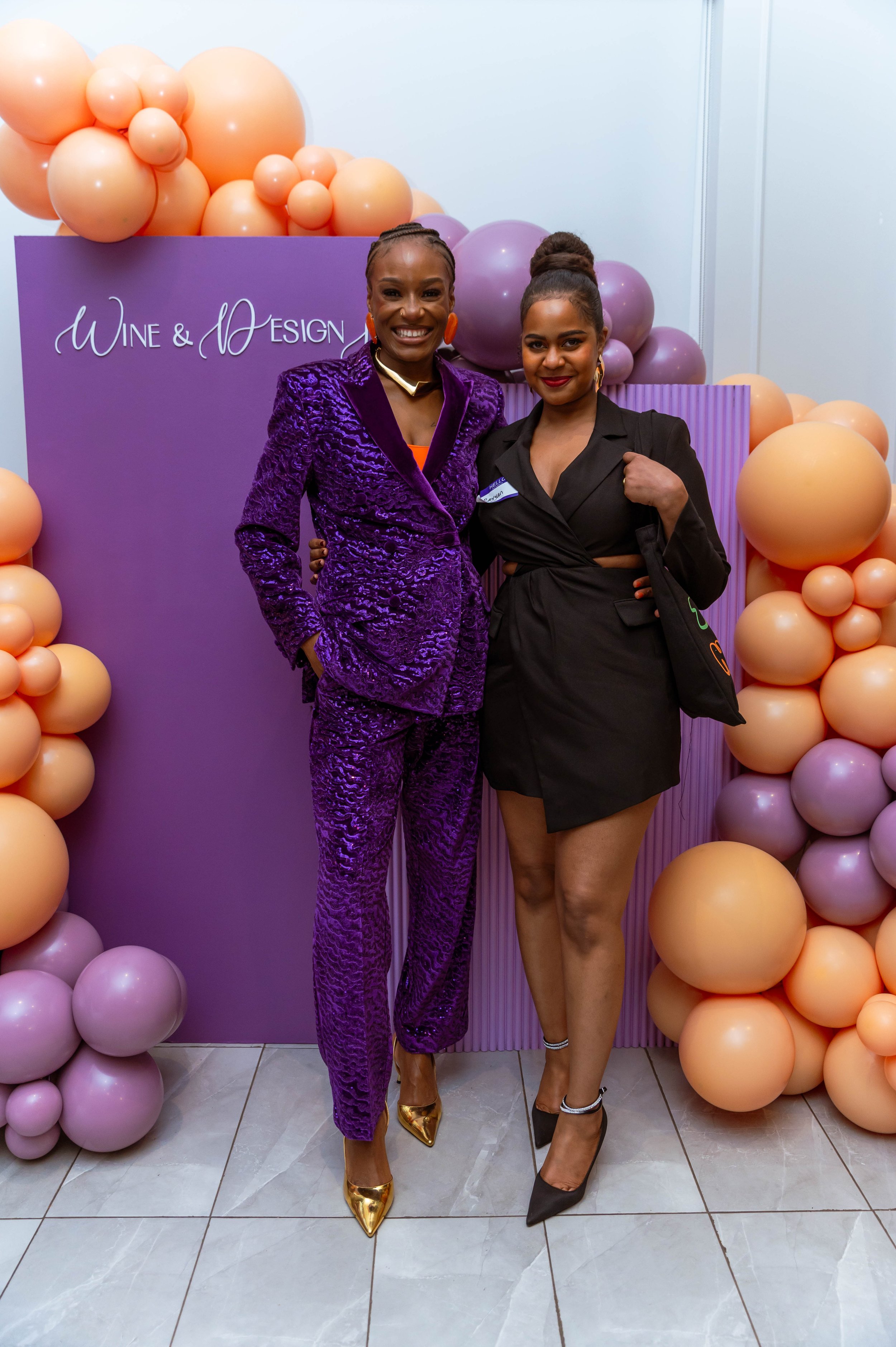 Two women posing together at an event with a purple backdrop and balloon decoration. One woman is wearing a purple textured suit with gold shoes, and the other is dressed in a black dress with high heels.