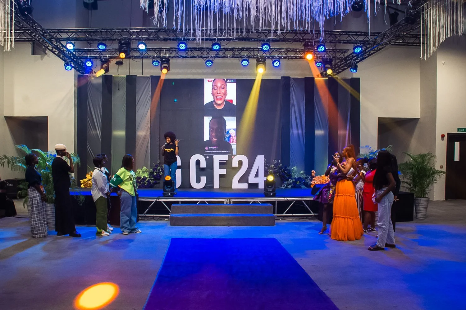 Group of people on a stage at an event, with a large digital screen showing two women, one smiling and one talking, and large letters reading 'ICF24'. Some attendees are taking photos, and the stage is decorated with plants and flowers under colorful