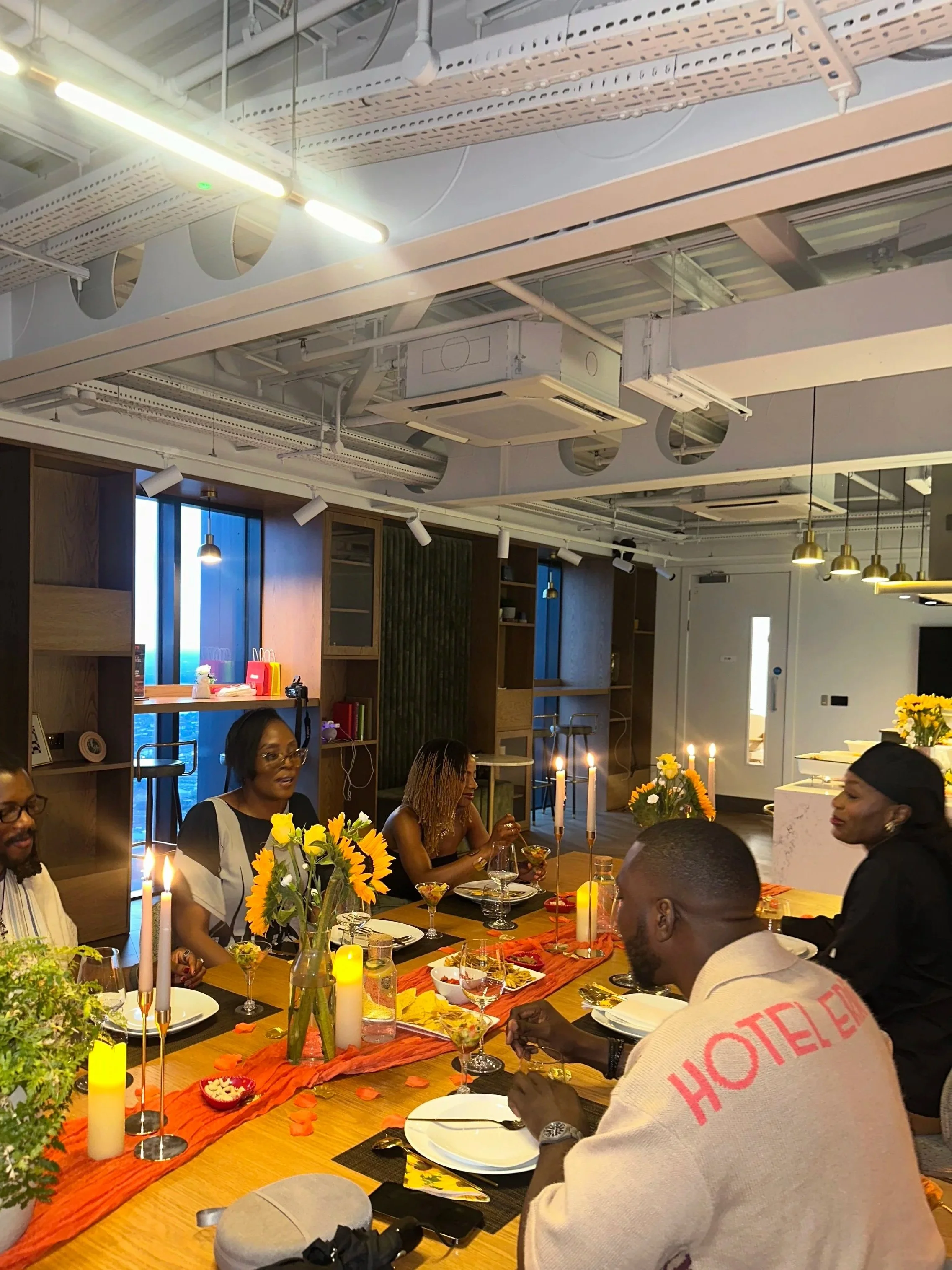 A group of people gathered around a dining table decorated with sunflowers, lit candles, and fall leaves, enjoying a meal together in a modern room with wooden shelving and hanging pendant lights.