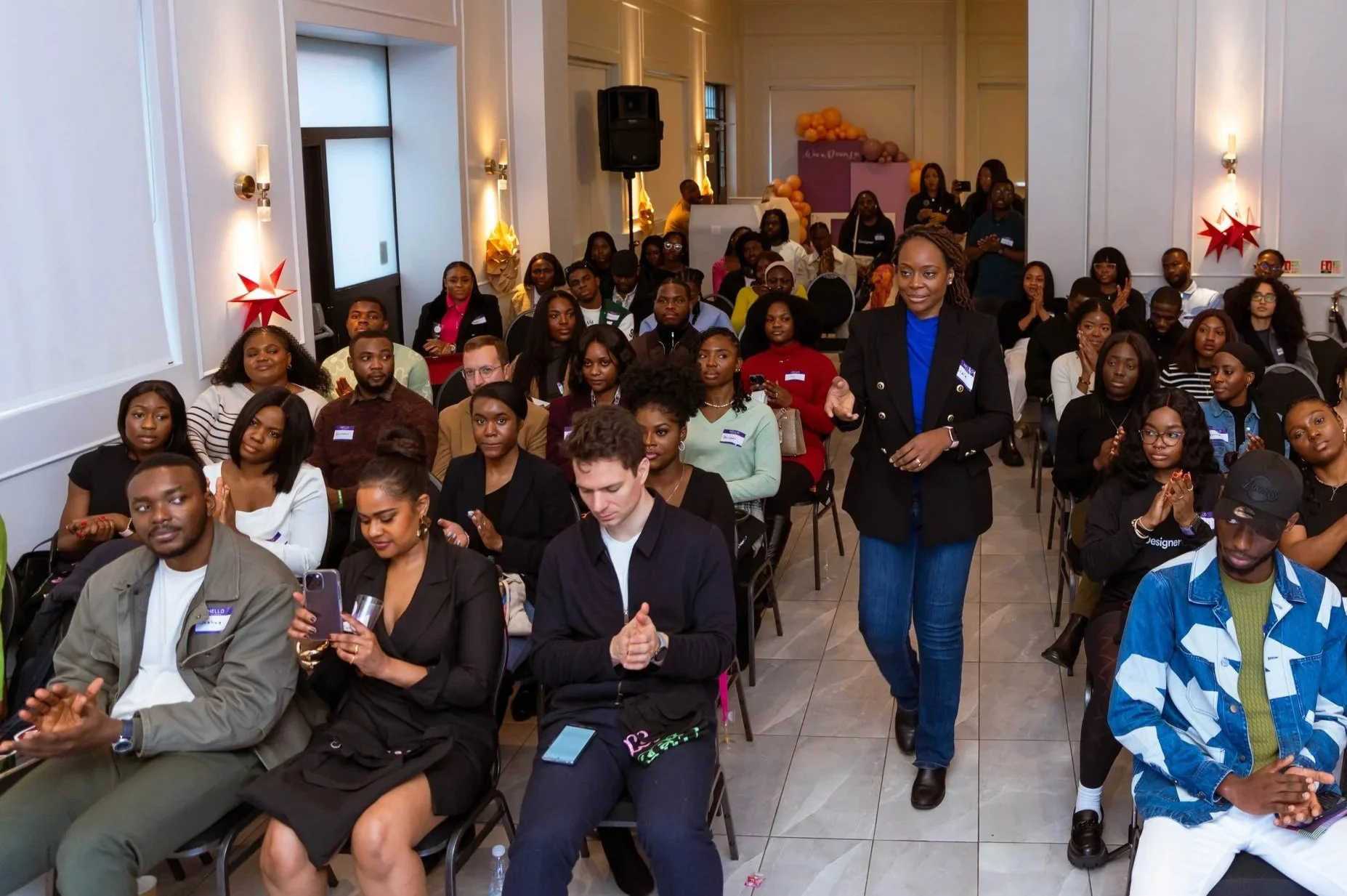 A diverse group of people attending a conference or seminar in a well-lit room with white walls and holiday decorations, with some people clapping and one woman standing and speaking.