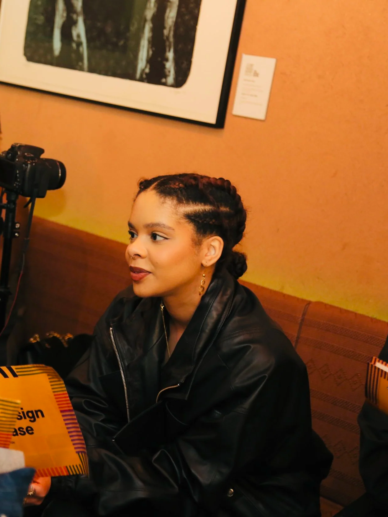A young woman with braided hair, wearing a black leather jacket, sitting indoors. There is a camera on a tripod and a colorful sign in front of her, with framed artwork on the wall behind her.