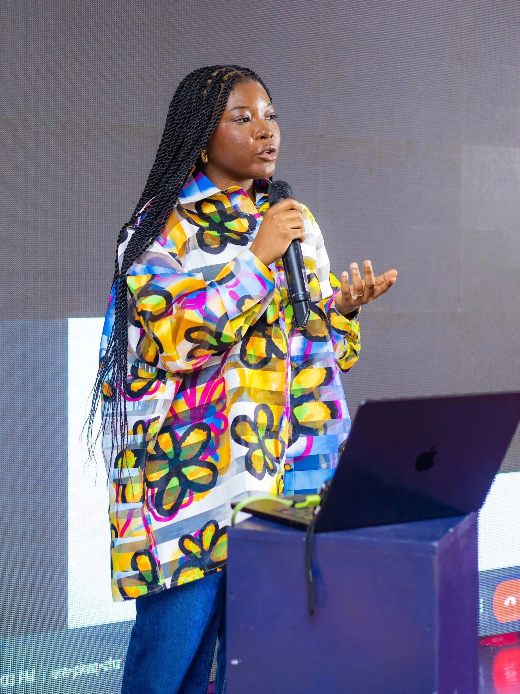 Woman speaking into microphone during presentation, standing behind a laptop on a table.
