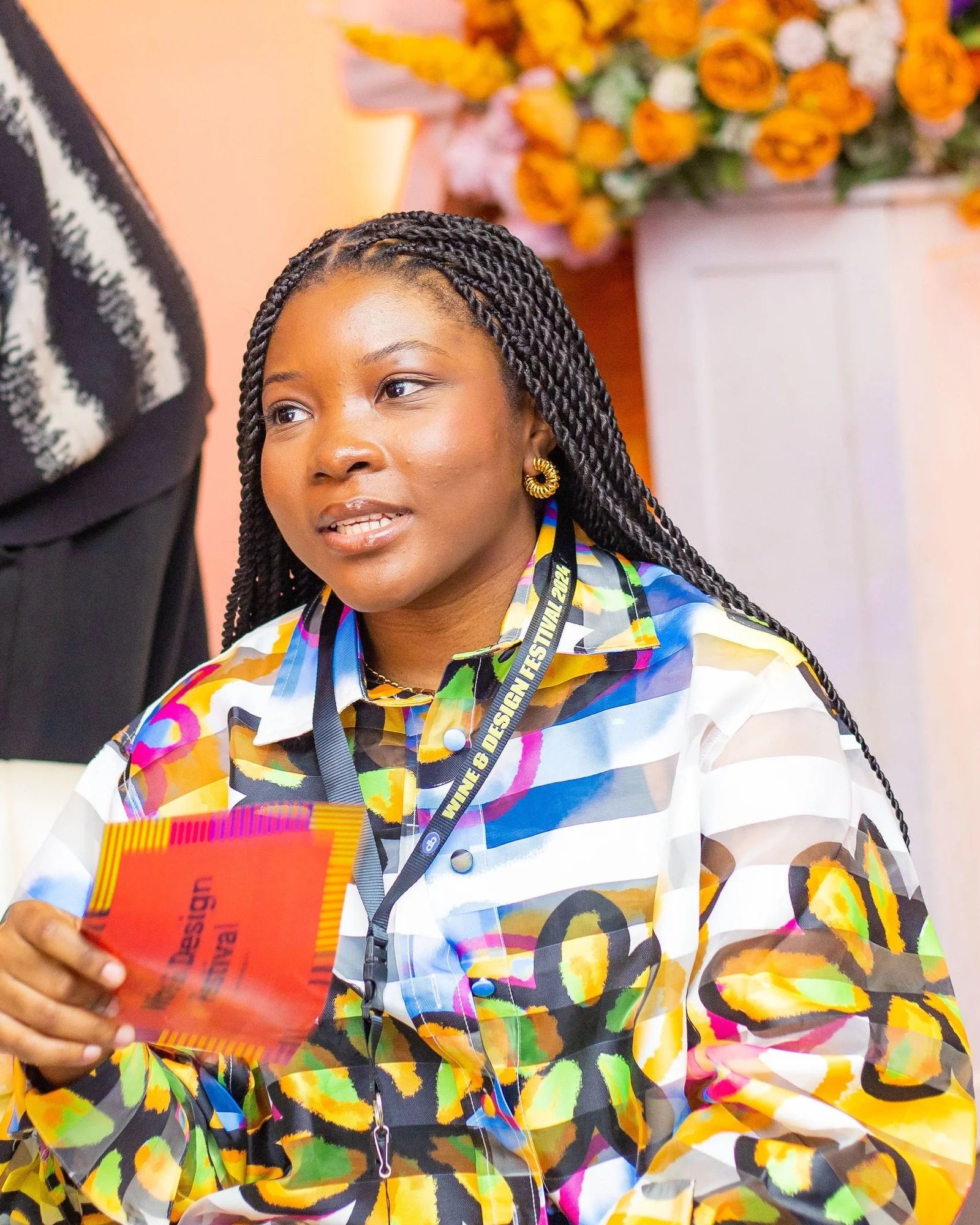 A woman with long braided hair wearing a colorful patterned blouse and gold jewelry, sitting indoor with a large floral arrangement in the background. She has a black lanyard around her neck and is holding a red book or folder.