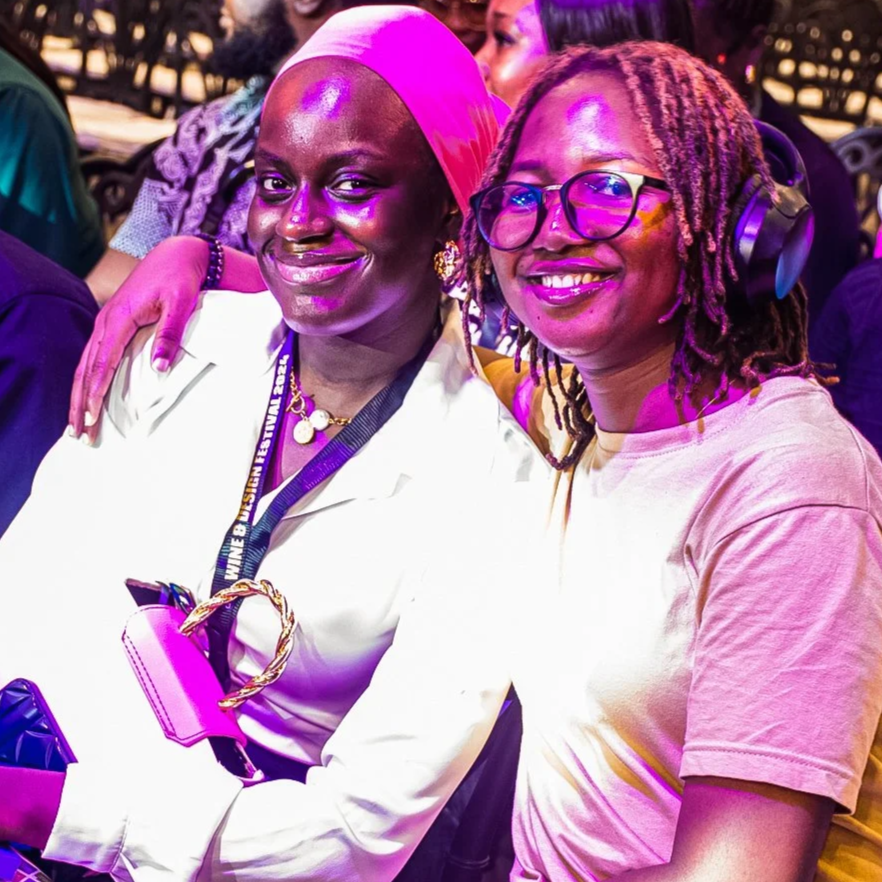 Two women smiling and sitting close together at an event, one with a pink headscarf and the other with glasses and dreadlocks, in a crowded venue with purple and yellow lighting.
