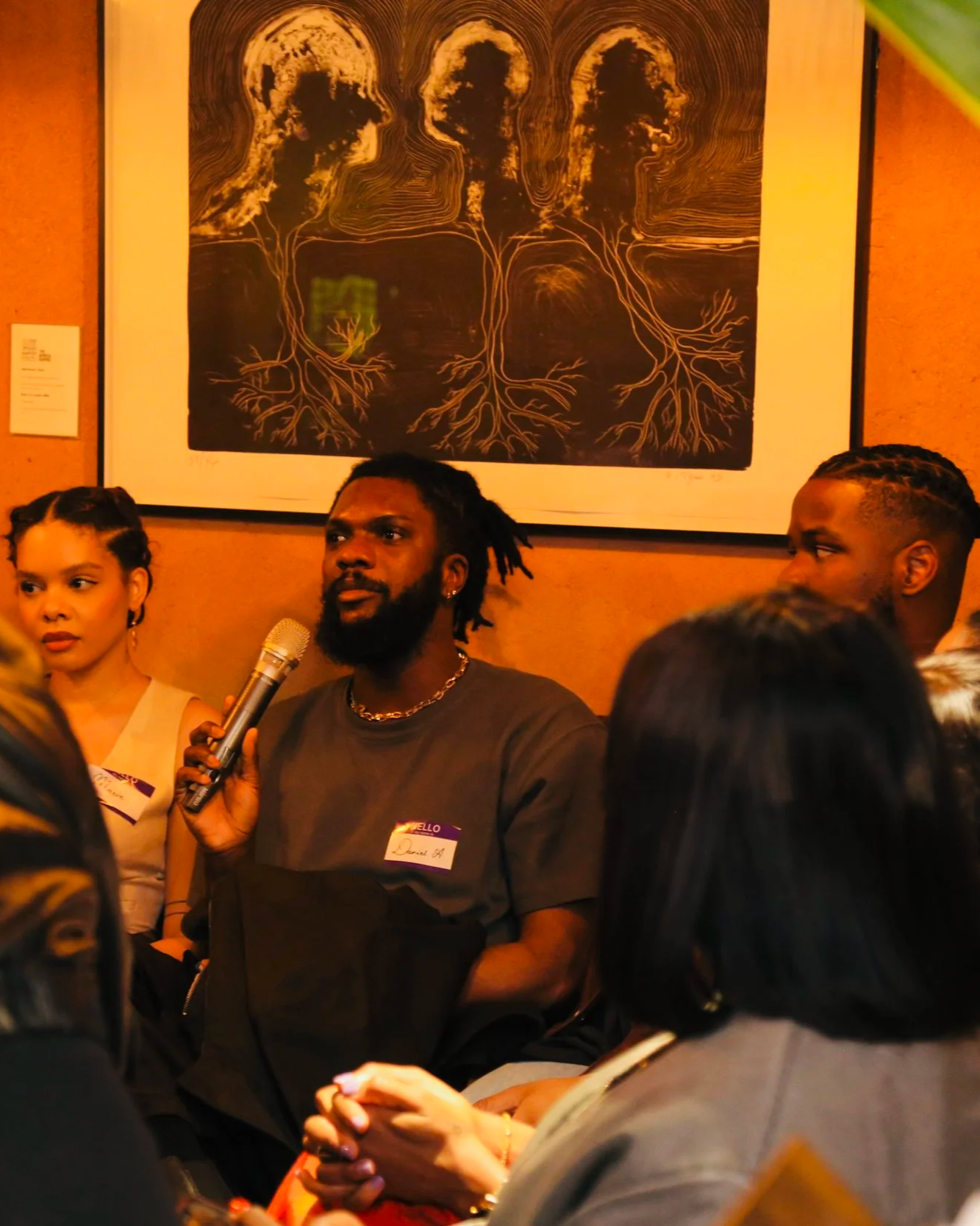 A group of people seated at a meeting or event, with a man holding a microphone and speaking. There is a framed artwork of three faces with tree roots depicted behind them on the wall.