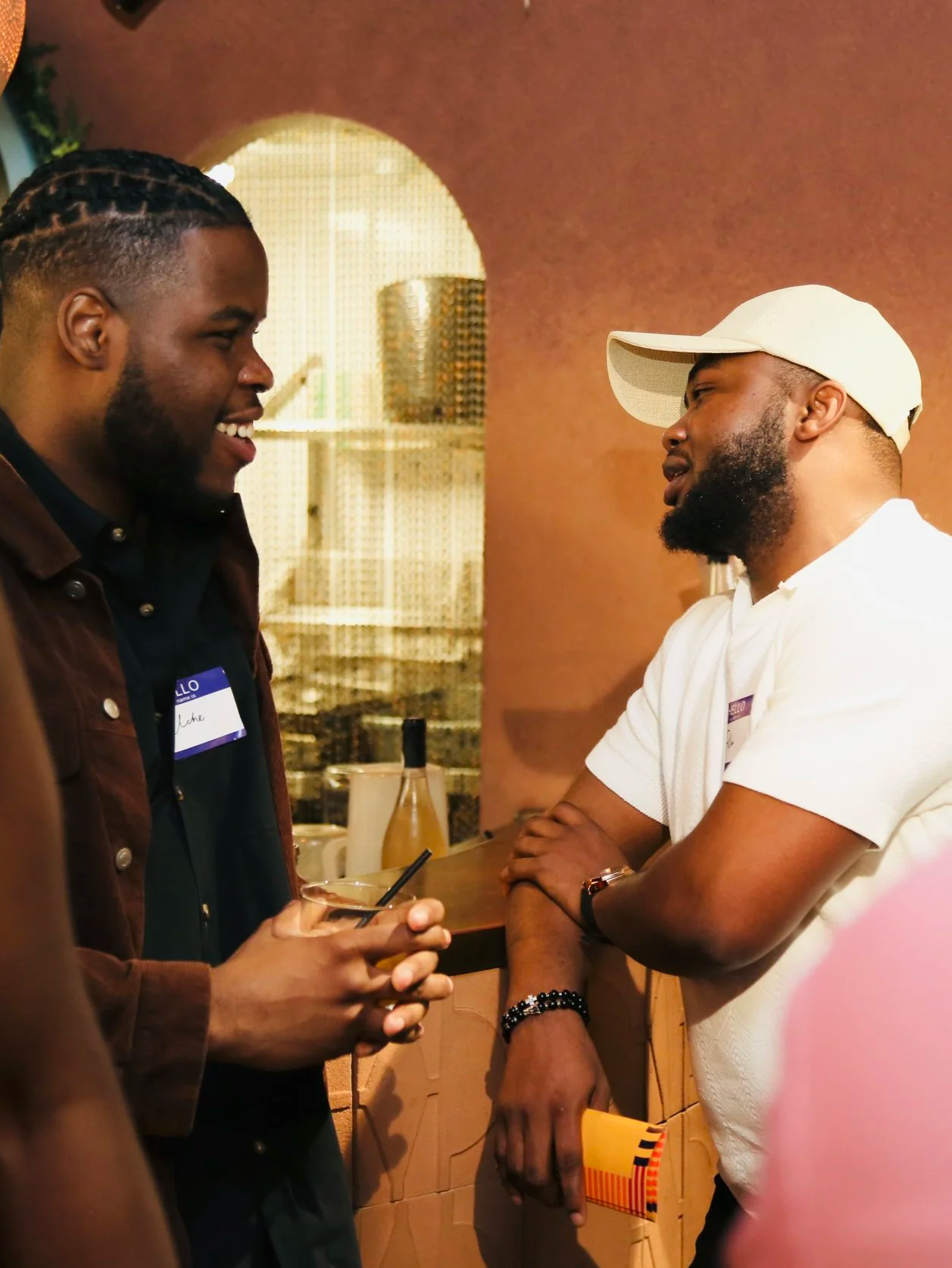 Two men engaged in conversation at a social gathering, one holding a drink with a straw, in a warmly lit room with a pink wall and decorative furniture in the background.