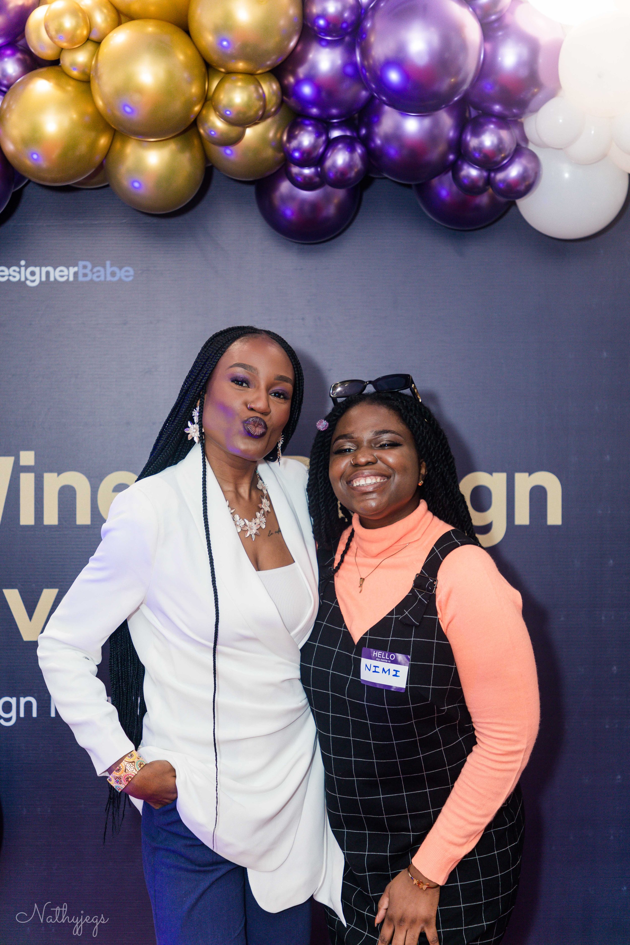 Two women smile and pose together at an event, standing in front of a dark backdrop with balloons in shades of gold, purple, and white. One woman wears a white blazer, blue pants, and purple lipstick, while the other wears a black plaid dress with a peach turtleneck and has sunglasses on her head.
