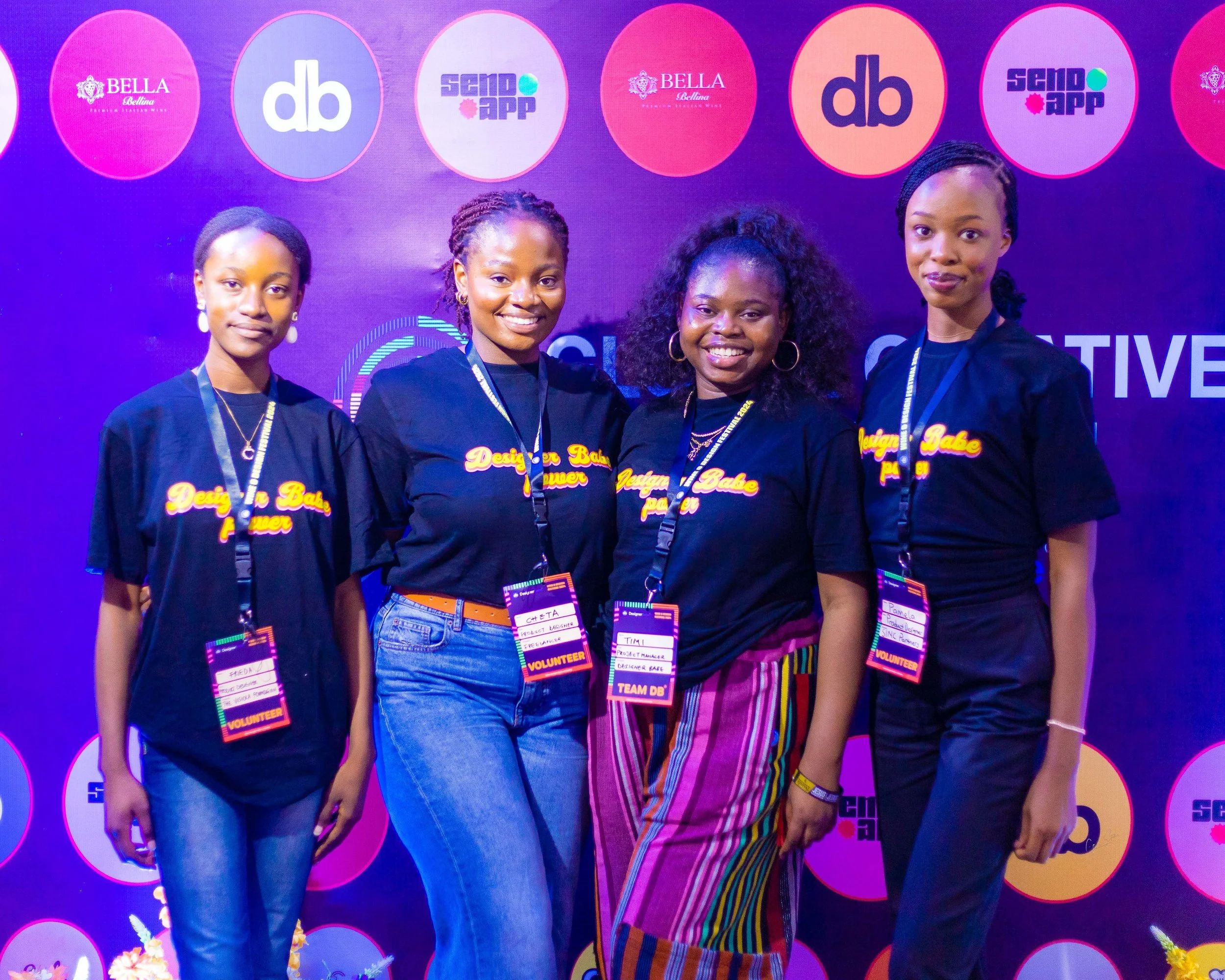 Four women standing together at an event, wearing black T-shirts with colorful text, each with a badge around their neck, against a backdrop with logos, smiling at the camera.