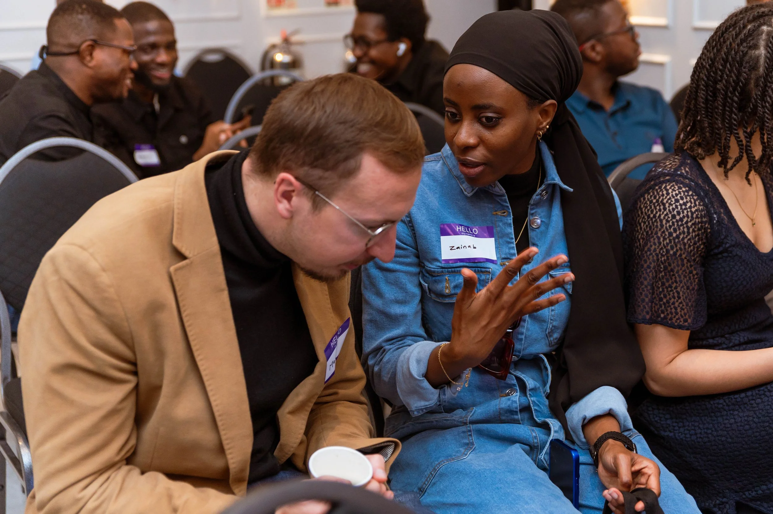 Two people sitting and engaged in conversation at a conference or seminar, with other attendees in the background.