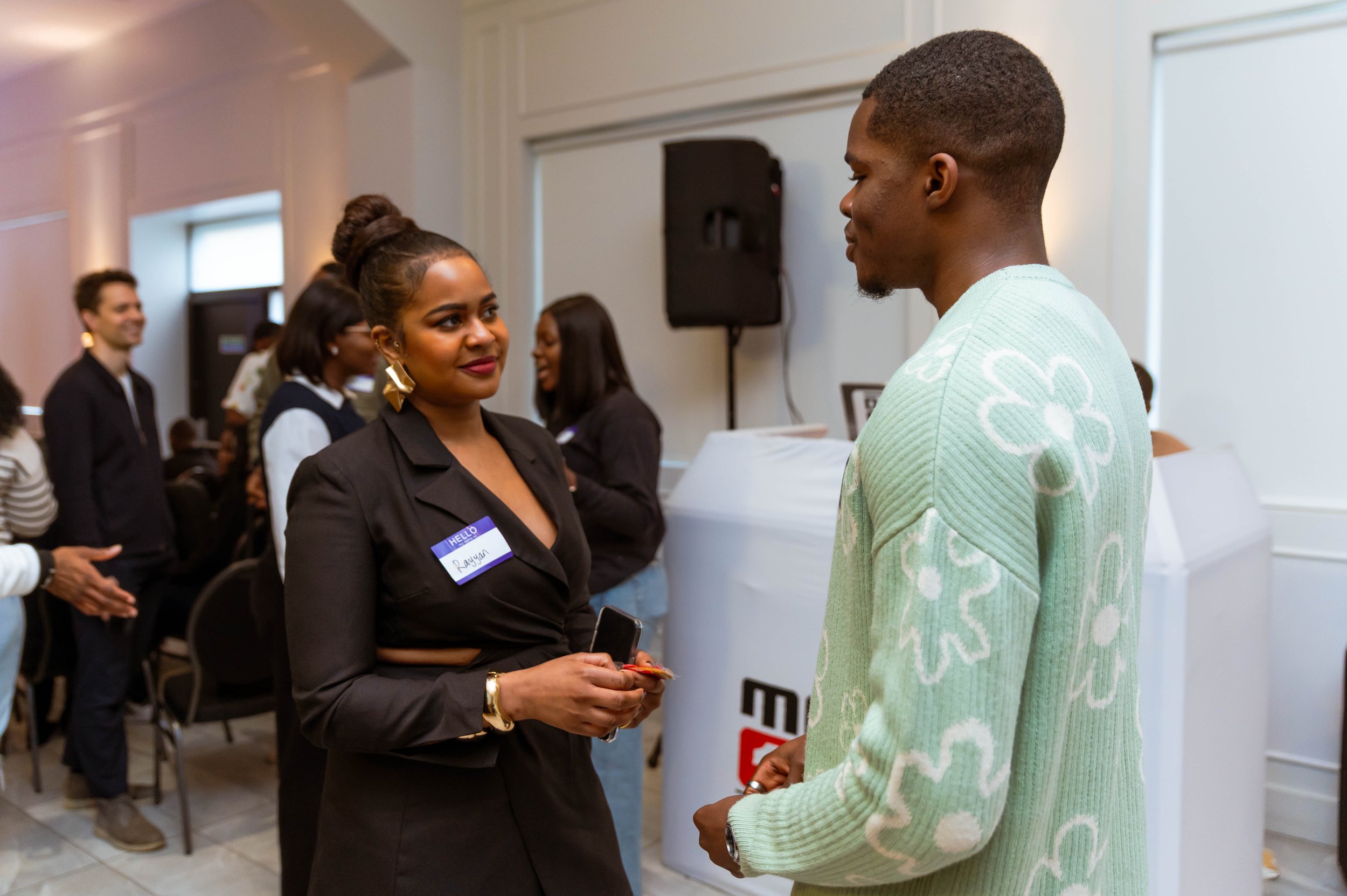 A woman and a man are engaged in conversation at a professional event. The woman is wearing a black blazer with a name tag, and the man is wearing a light green sweater with white patterns. In the background, other attendees are socializing in a well-lit room.