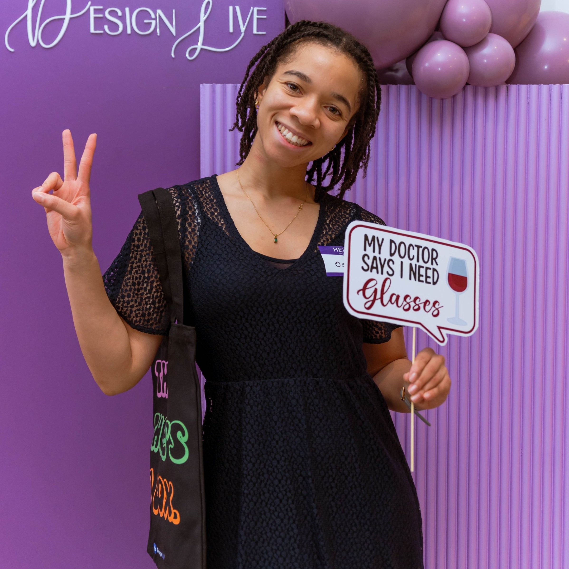 A smiling woman with braids holding a sign that reads "My doctor says I need glasses" and making a peace sign with her other hand, standing in front of a purple backdrop with balloons and part of a sign visible.