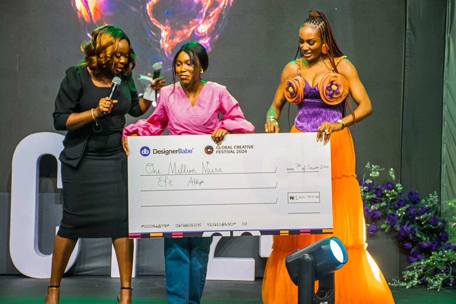 Three women on a stage holding a large check, with purple flowers and stage lighting in the background. The check is from the Global Creative Festival 2024 and is made out to 'One Million Naira' for Efie Abena. The woman in the middle is wearing a pi