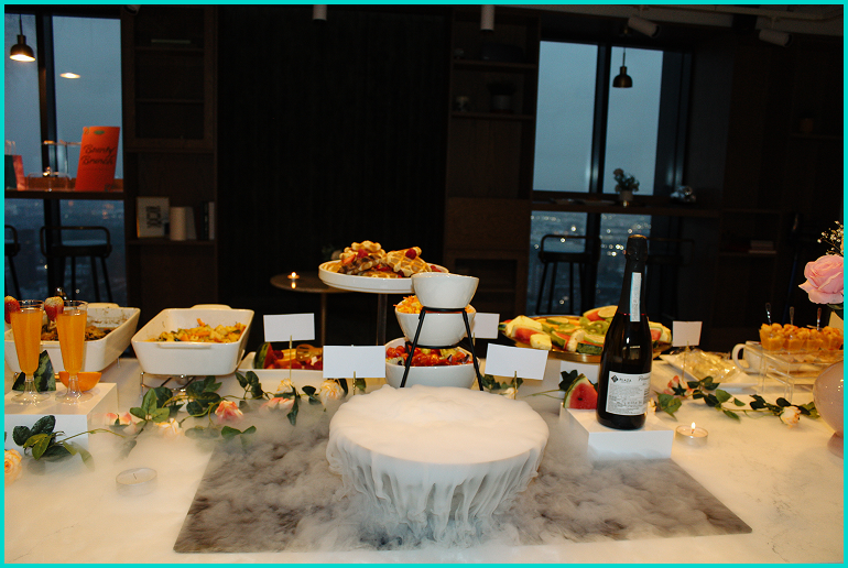 Buffet table with a dry ice centerpiece, various food dishes, a bottle of wine, and decorative flowers in an indoor setting with large windows.