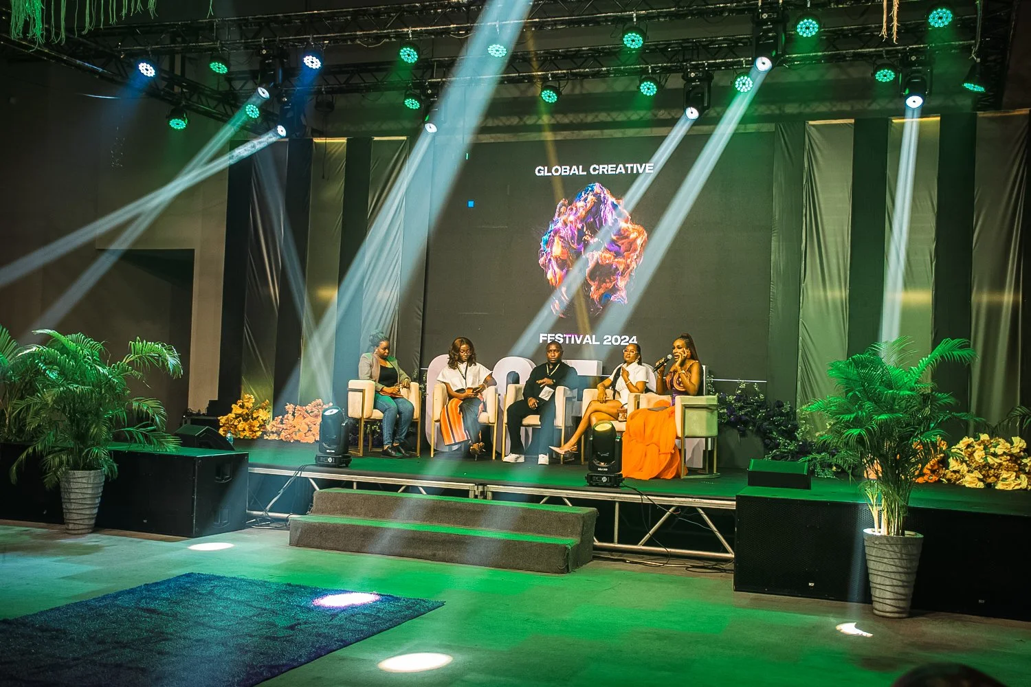 A panel discussion on stage at a festival with five people seated in chairs. The background displays a large screen with the words 'Global Creative Festival 2024' and a colorful abstract graphic. The stage is decorated with potted plants and flowers,