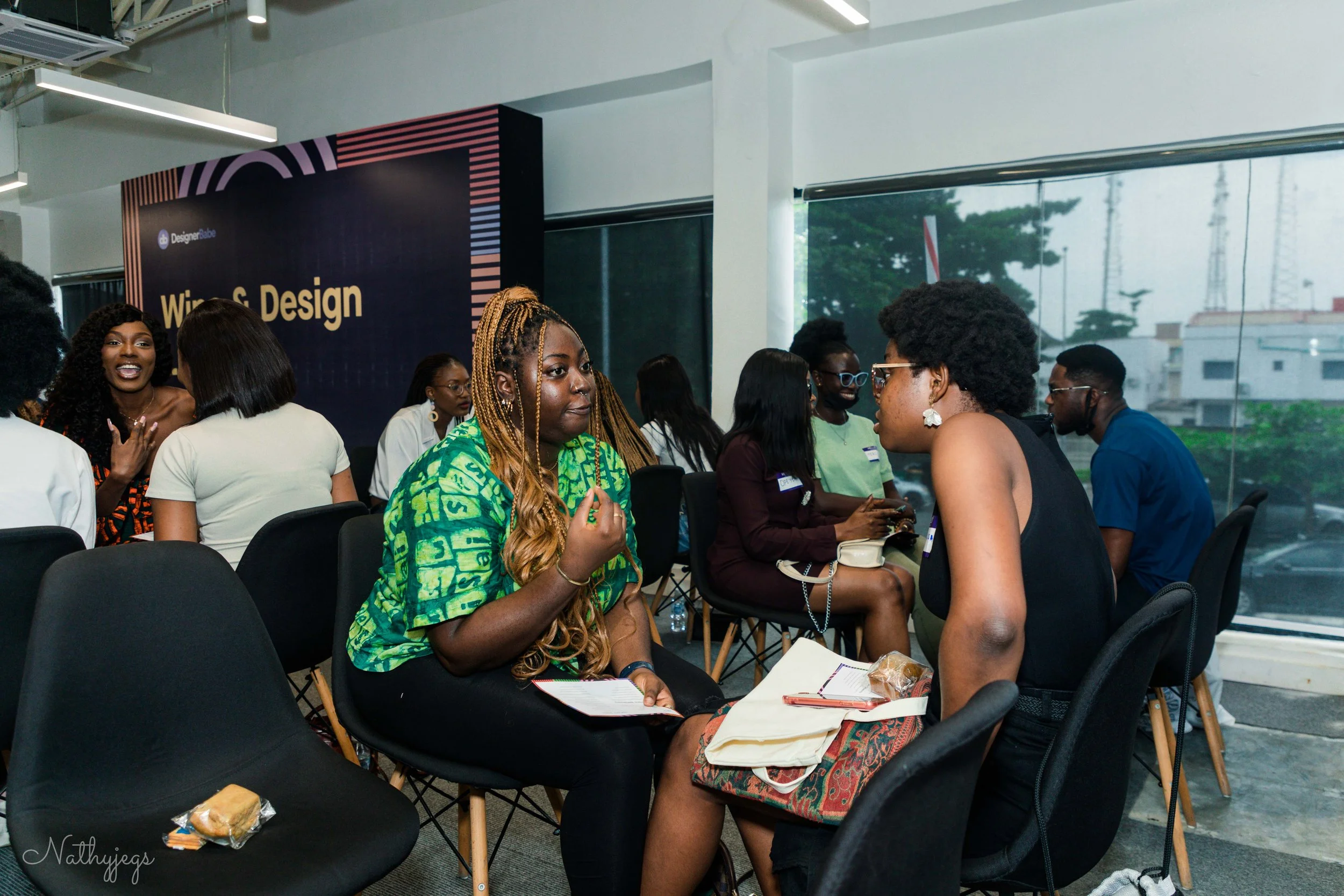 Women sitting and conversing at a workshop with a sign that reads "Wine & Design" in the background.
