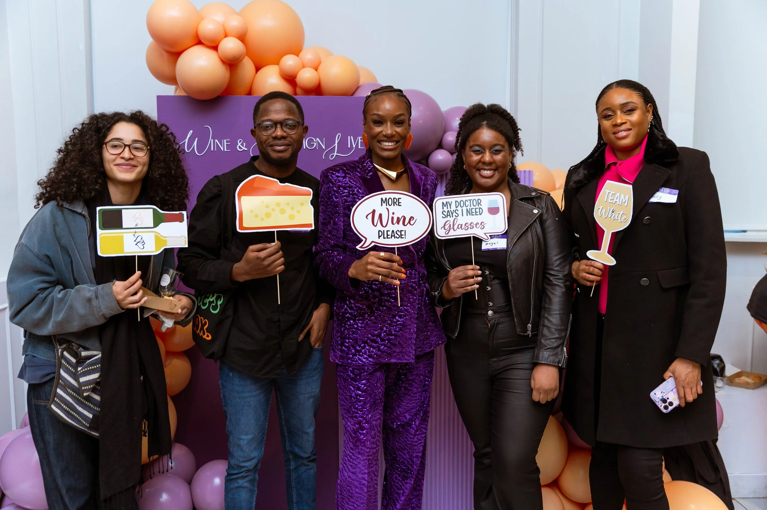 Group of five diverse people smiling at a wine and design event, holding playful signs with wine-related messages, standing in front of balloons and a purple backdrop.