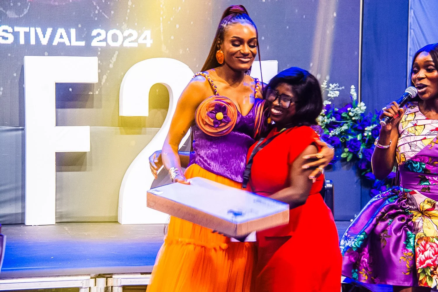 Three women celebrating at an event, one holding a box, with a backdrop displaying 'Festival 2024' and large initials 'EG'.