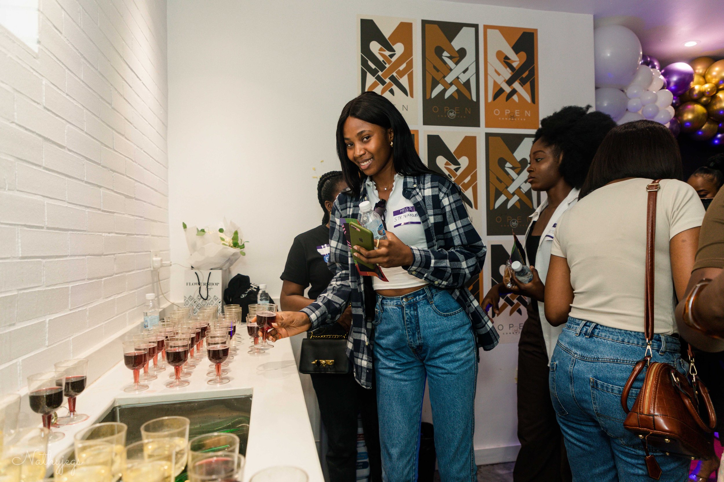 Women at a gathering with drinks, one woman in the foreground holding a drink and a bottle, surrounded by others, in a room with white brick wall, posters, and balloons.