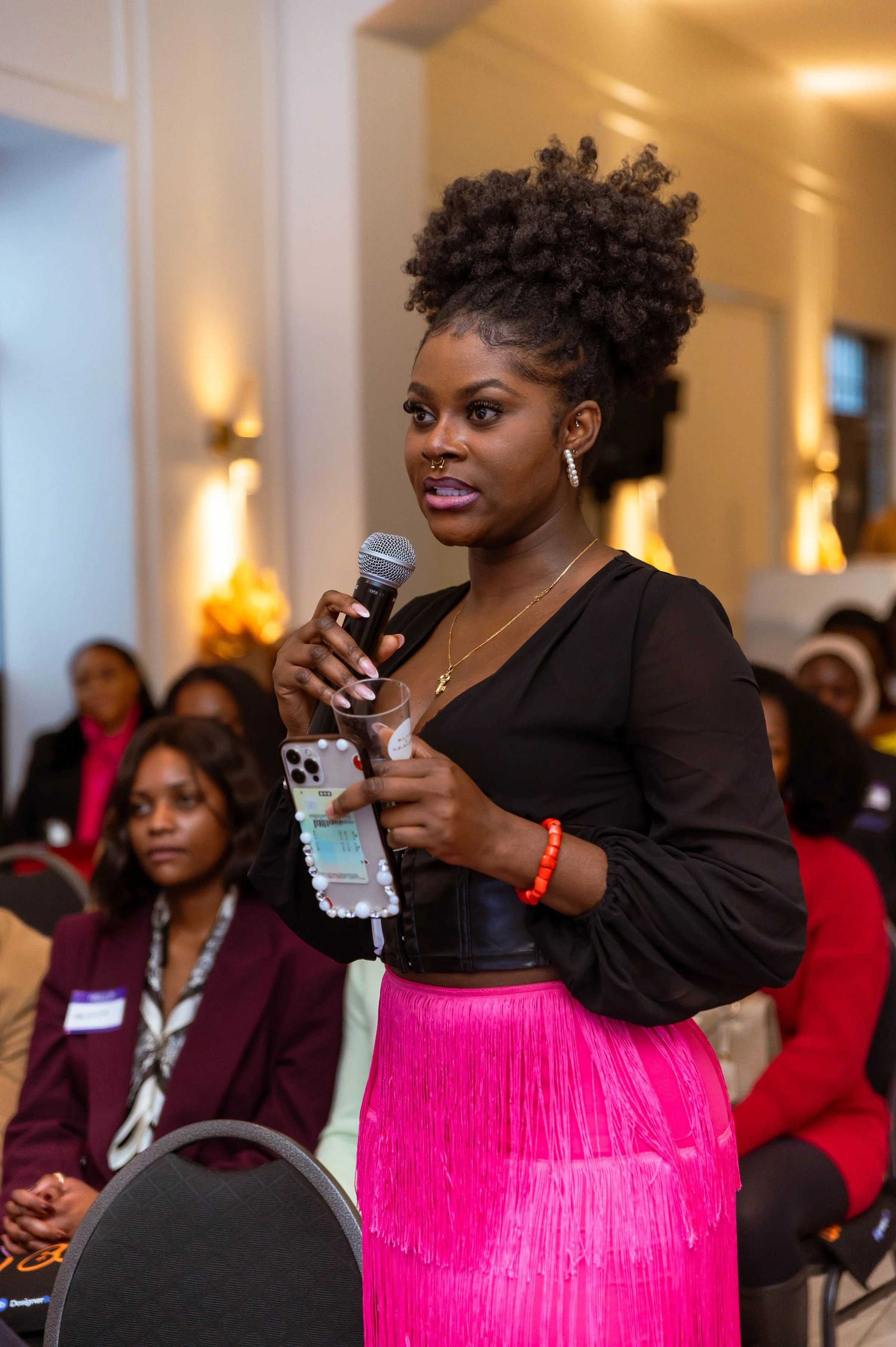 A woman with curly hair, earrings, and a necklace is speaking into a microphone at a formal event. She is wearing a black top and a bright pink skirt, holding a glass and a smartphone. Several people are seated behind her, listening attentively.