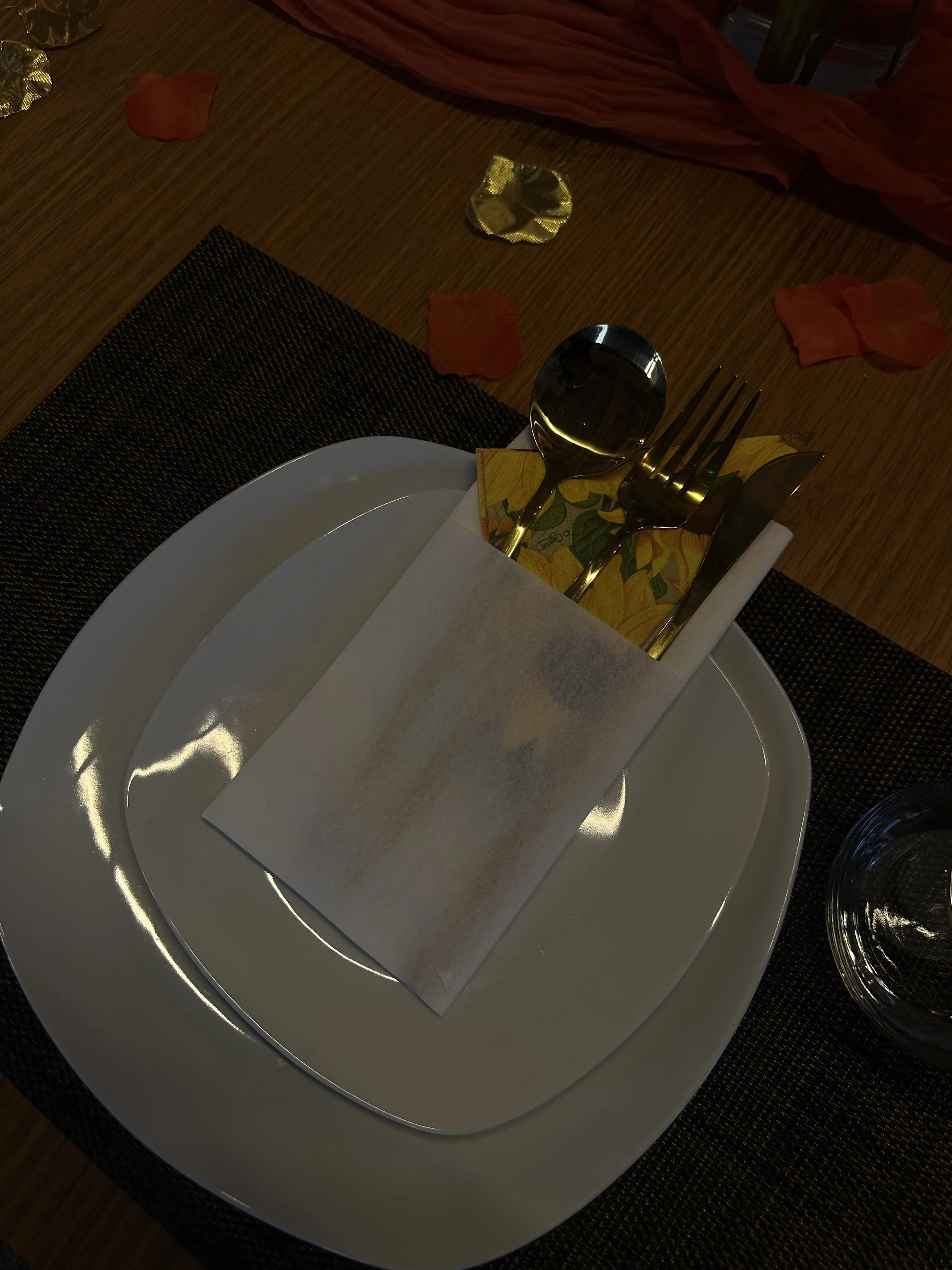 Set dining table with a white plate, a fork, a knife, a spoon, a cloth napkin, and a paper holder with a floral design, on a dark placemat, with scattered flower petals on a wooden table.