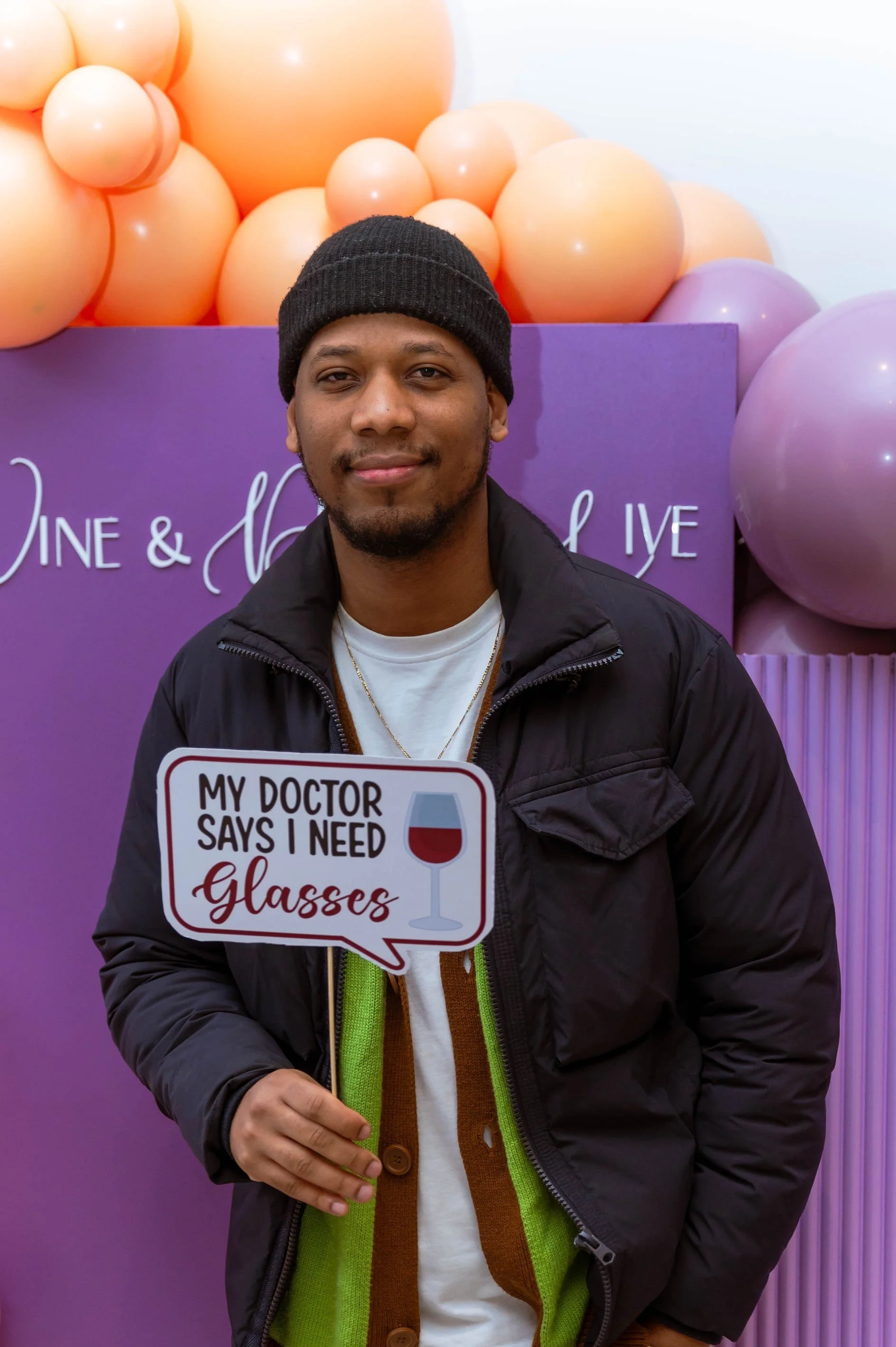 A young man with a black beanie and black jacket, smiling, holding a sign that reads 'My doctor says I need glasses' with a graphic of a wine glass. Background includes purple balloons and a purple sign with cursive writing.