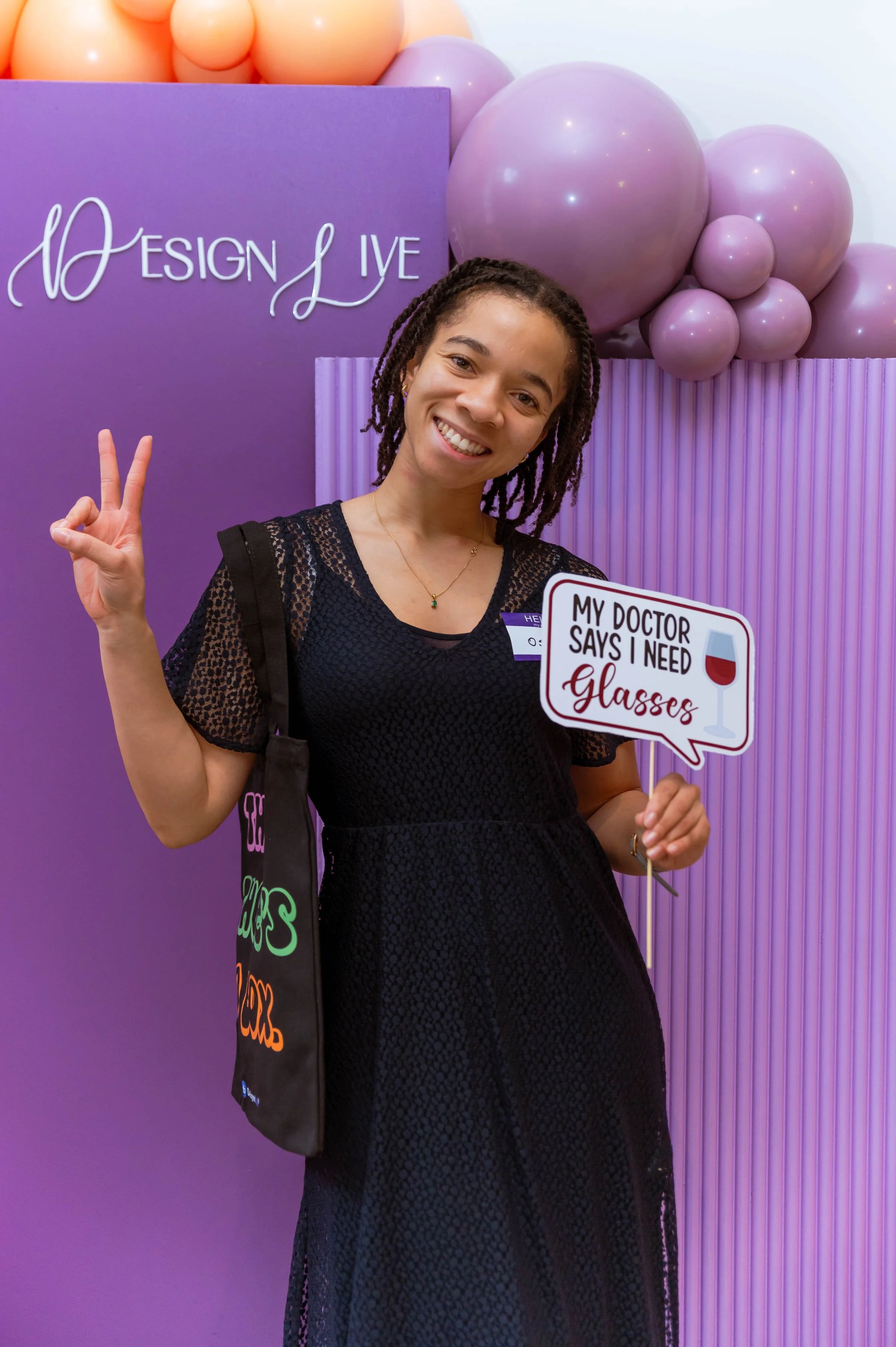 Smiling woman holding a sign that reads "My doctor says I need glasses" at an event with purple balloons and a purple background that says "Design Live."