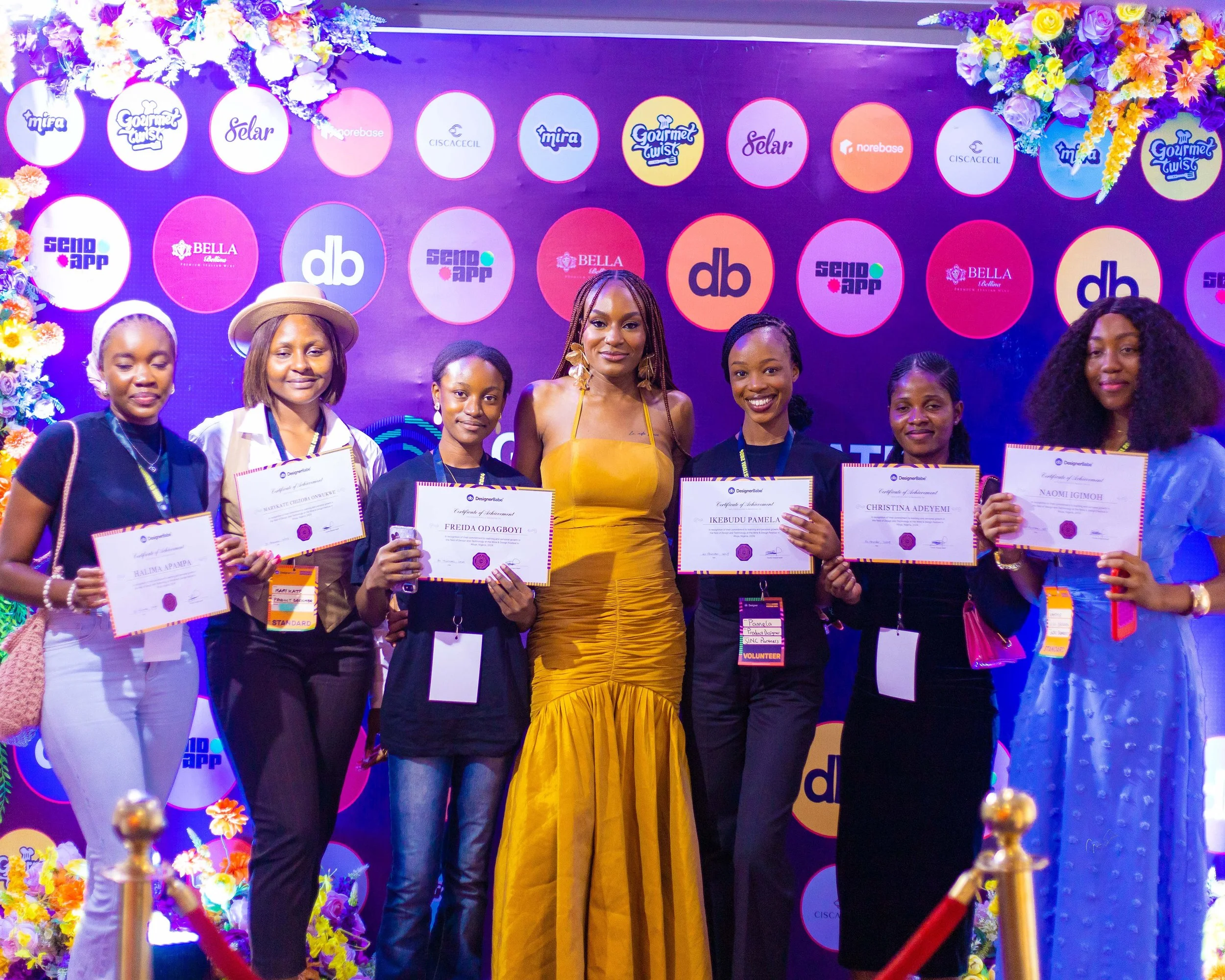 A group of seven women standing on a stage, holding certificates at an event with a colorful backdrop featuring various logos and floral decorations.