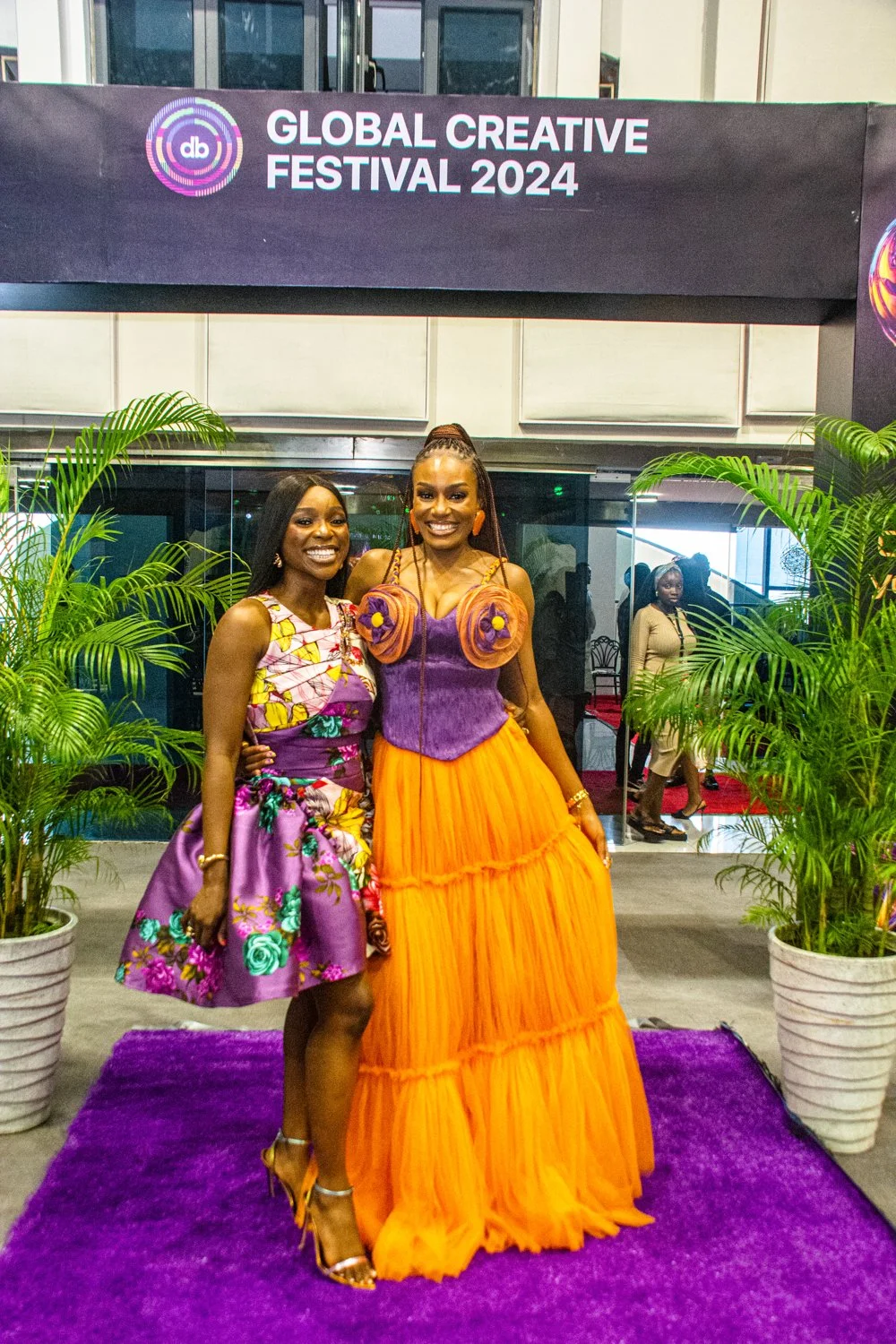 Designer Babe and Tomike Adeoye in vibrant, colorful dresses pose on a purple carpet at the Global Creative Festival 2024, with green plants on either side and a banner overhead.