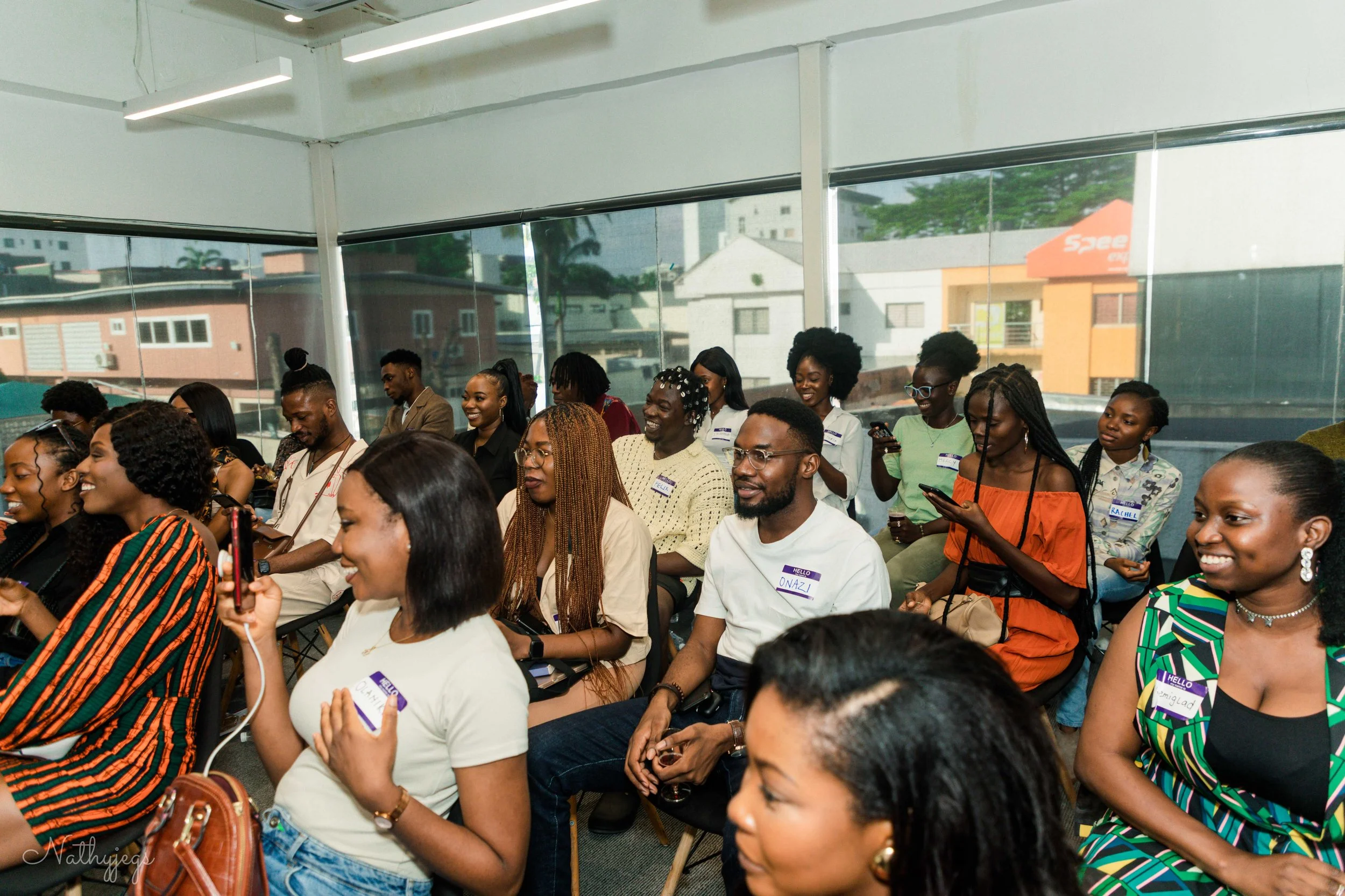 Group of diverse people seated and smiling during an indoor event, some wear name tags, while a few are using phones.