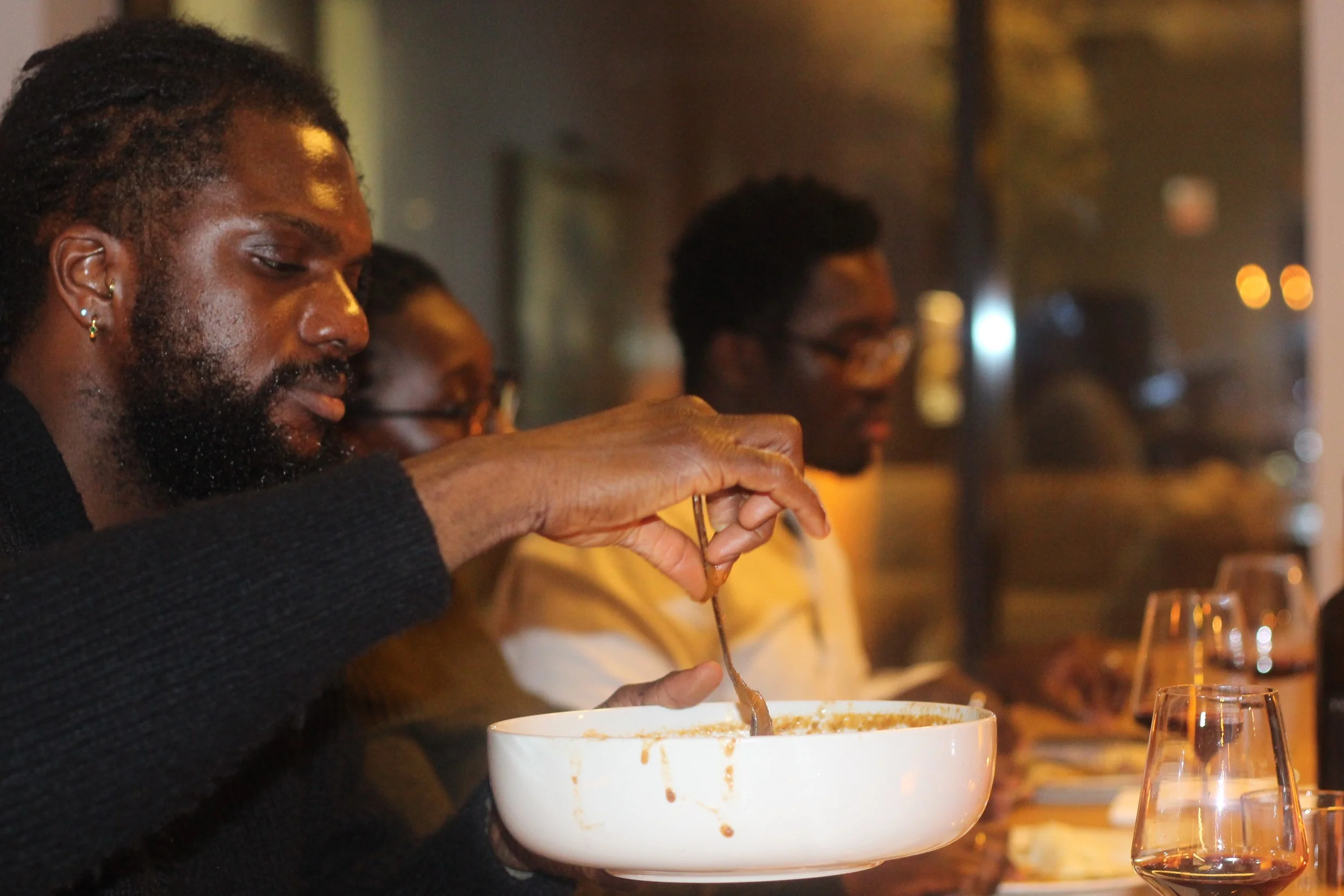 Man with a beard and earrings scooping food from a large white bowl at a dinner table, with two people in the background.
