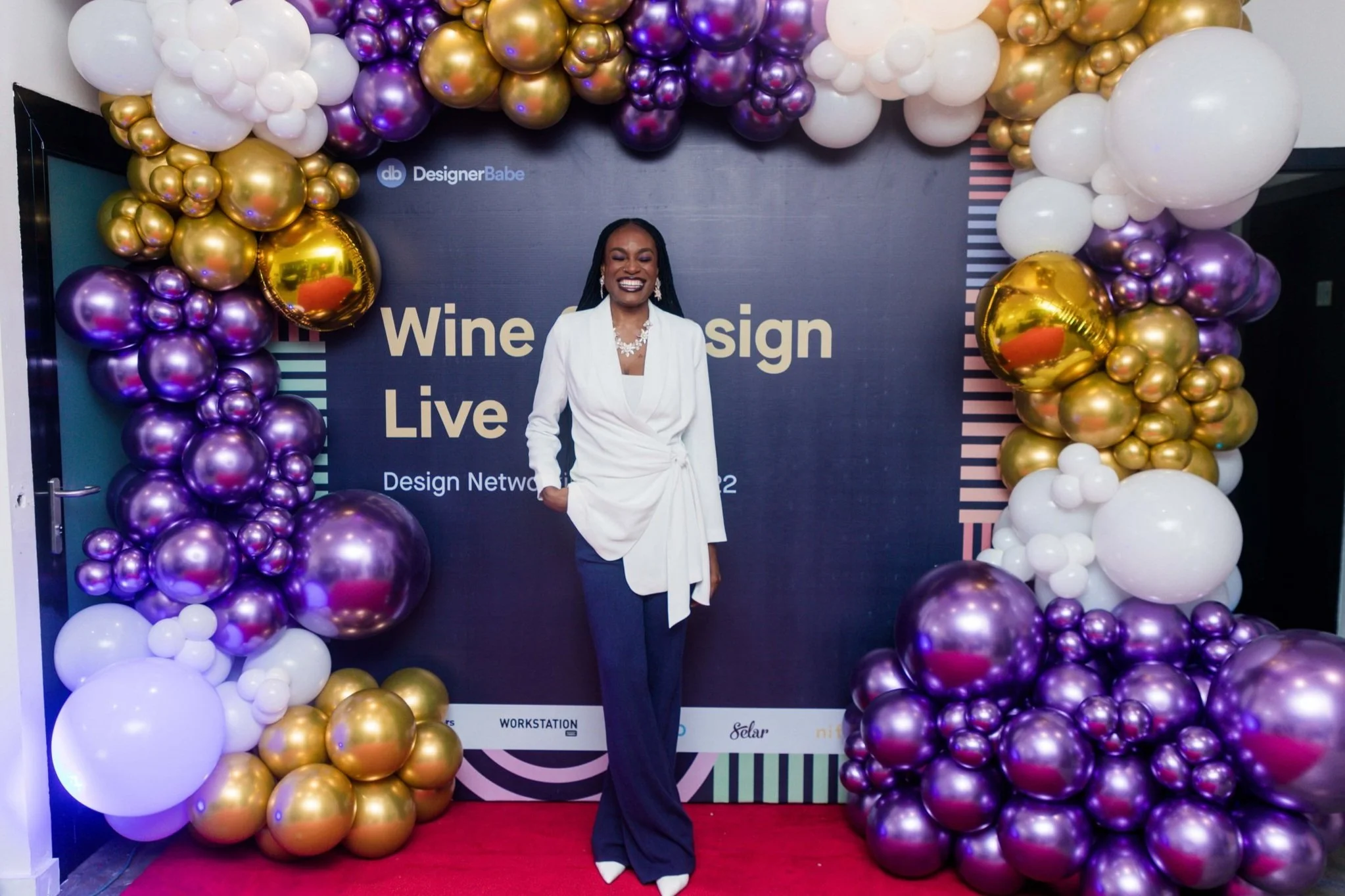 A woman in a white blazer and dark pants standing in front of a decorated backdrop with multicolored balloons in purple, gold, and white, at a wine design live event.
