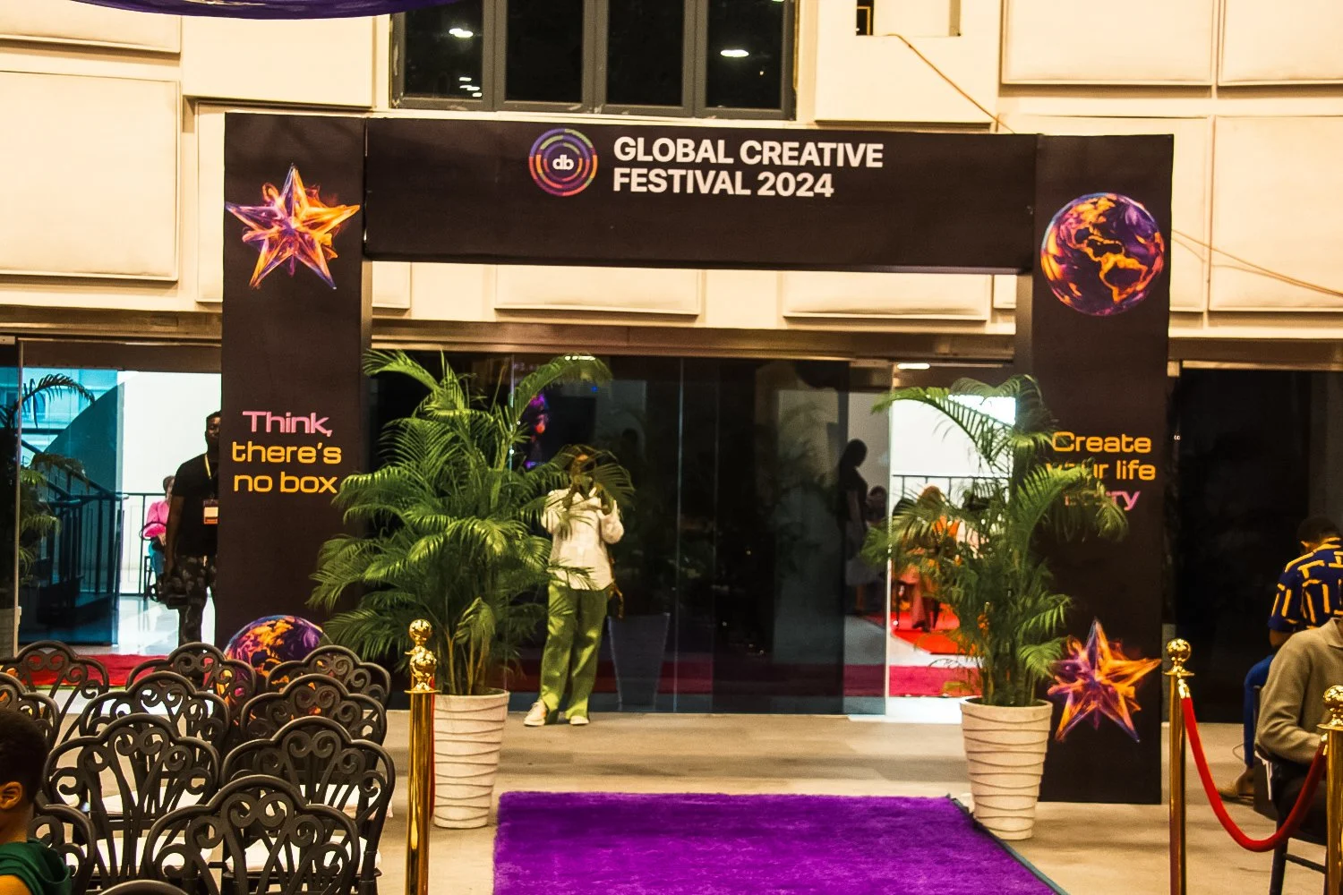 Entrance to the Global Creative Festival 2024, decorated with potted plants, colorful logo symbols, black banners with text, and a purple carpet leading into the event area.