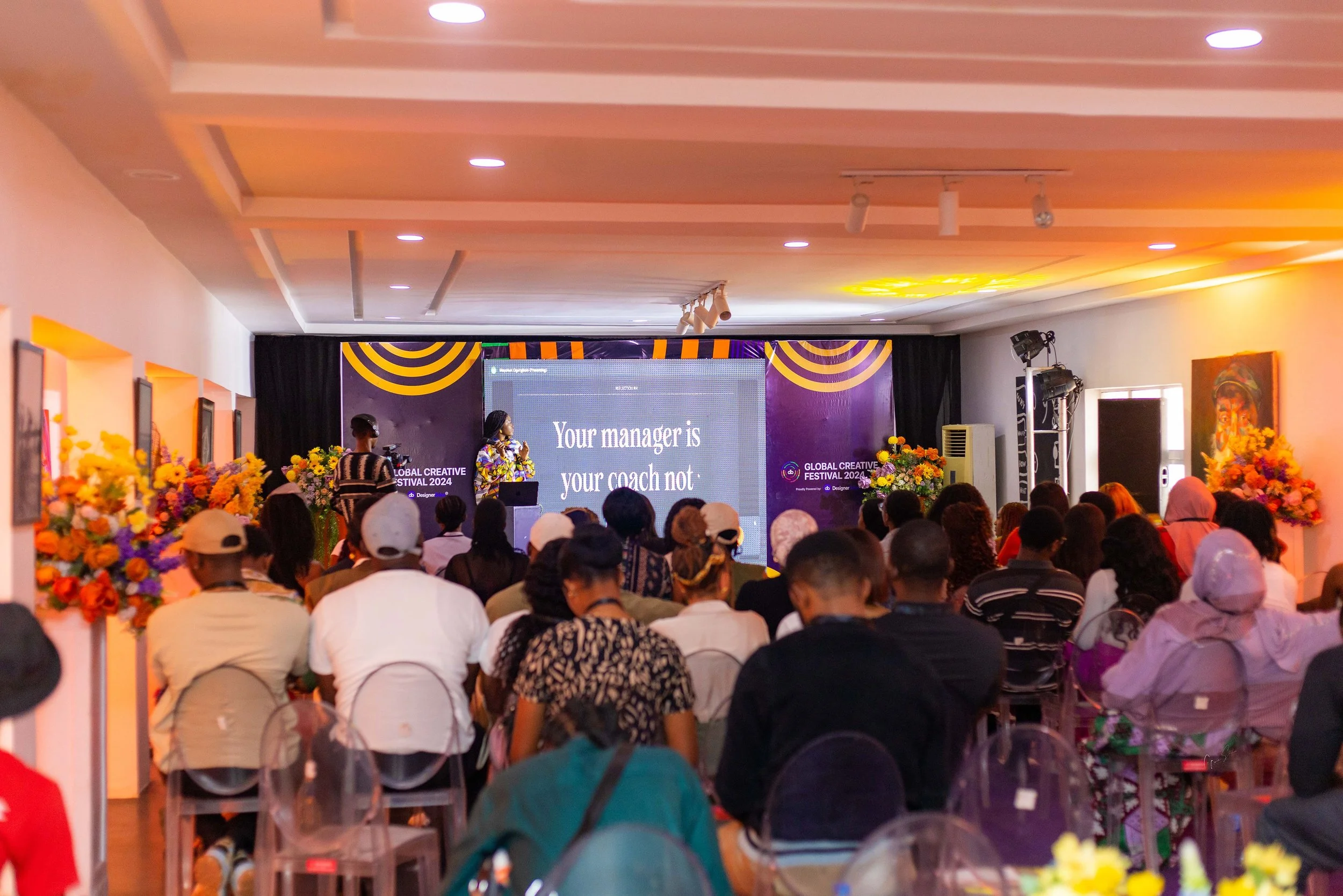 A large group of people seated and attending a presentation at the Global Creative Festival 2024, with a woman speaking on stage and a large screen displaying the text, 'Your manager is your coach not:'.