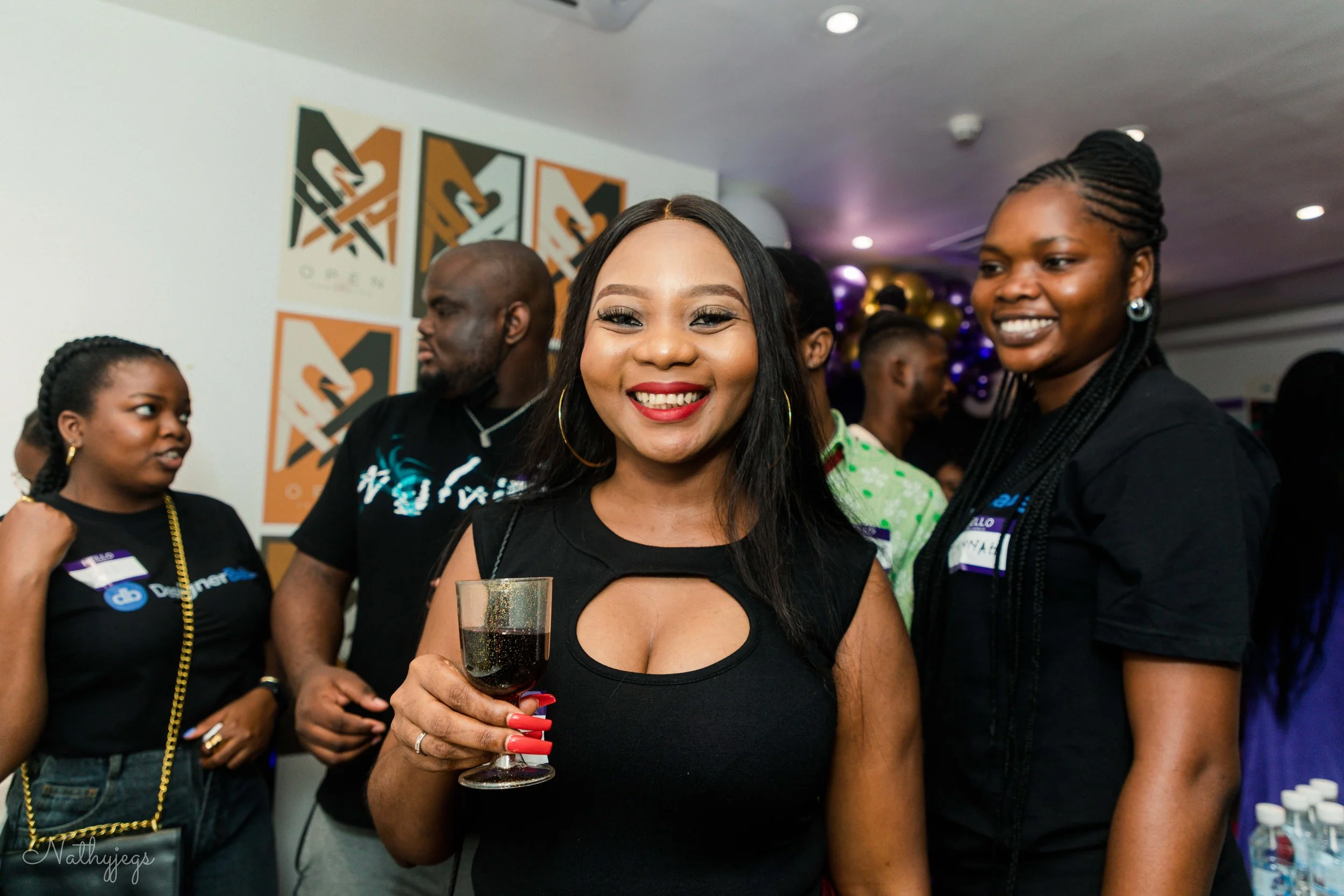 Smiling woman with dark hair and red lipstick holding a glass of red wine at a party, surrounded by people in casual attire, with colorful wall art and balloons in the background.